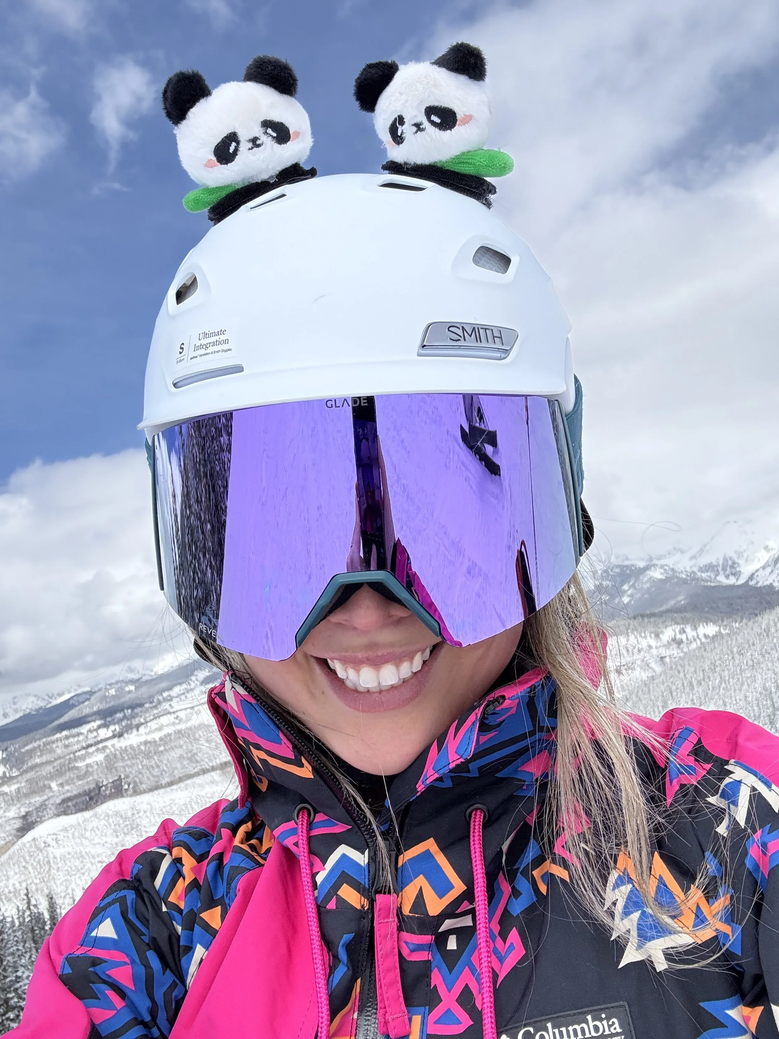 Person wearing a ski helmet with panda plush toys on top, reflective ski goggles, and a colorful Columbia winter jacket, smiling in a snowy mountain landscape.