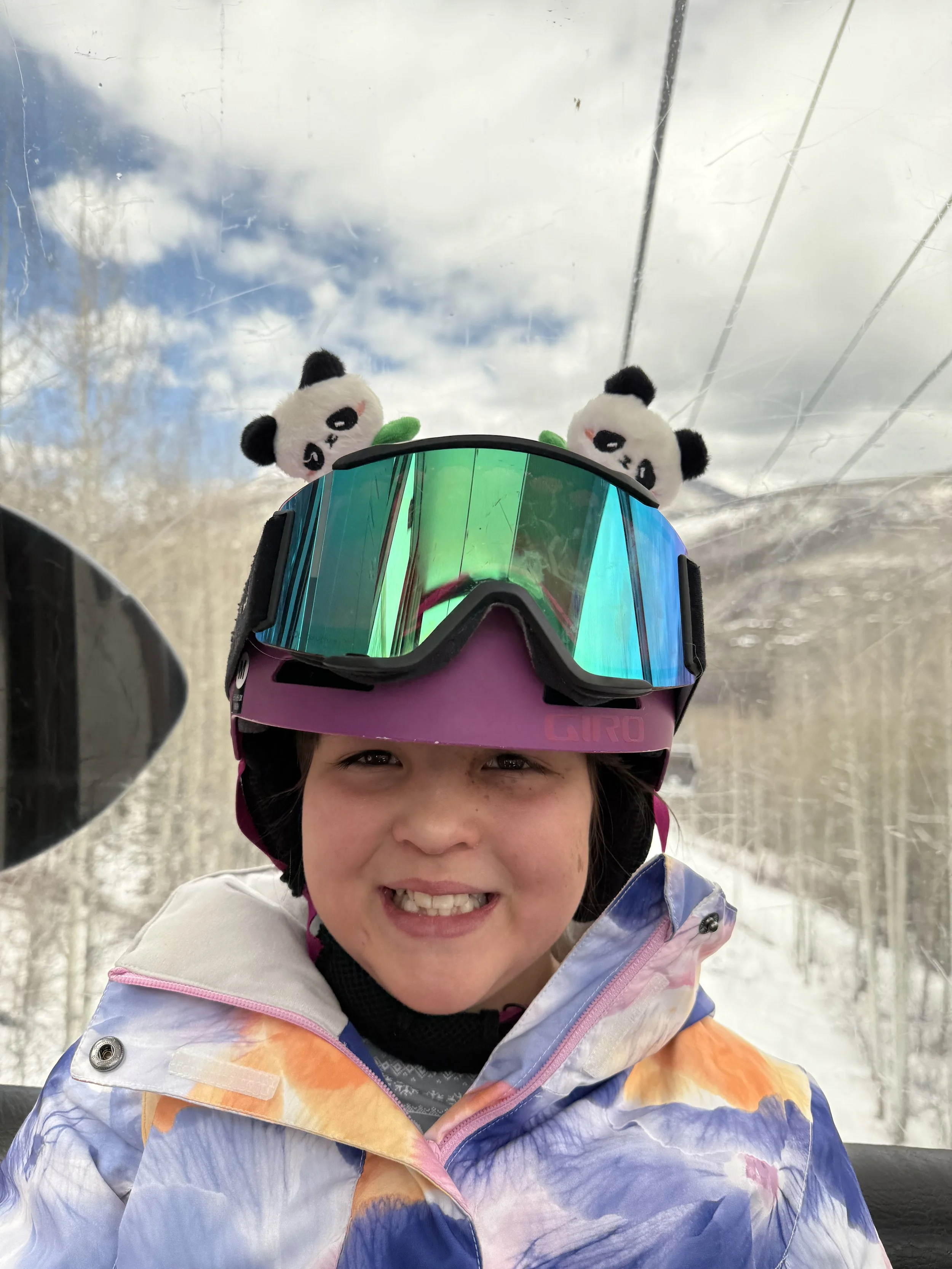 A young girl in winter gear with a colorful jacket, wearing a pink ski helmet with panda plush toys on top, and reflective ski goggles, smiling inside a ski lift with snowy trees and cloudy sky in the background.