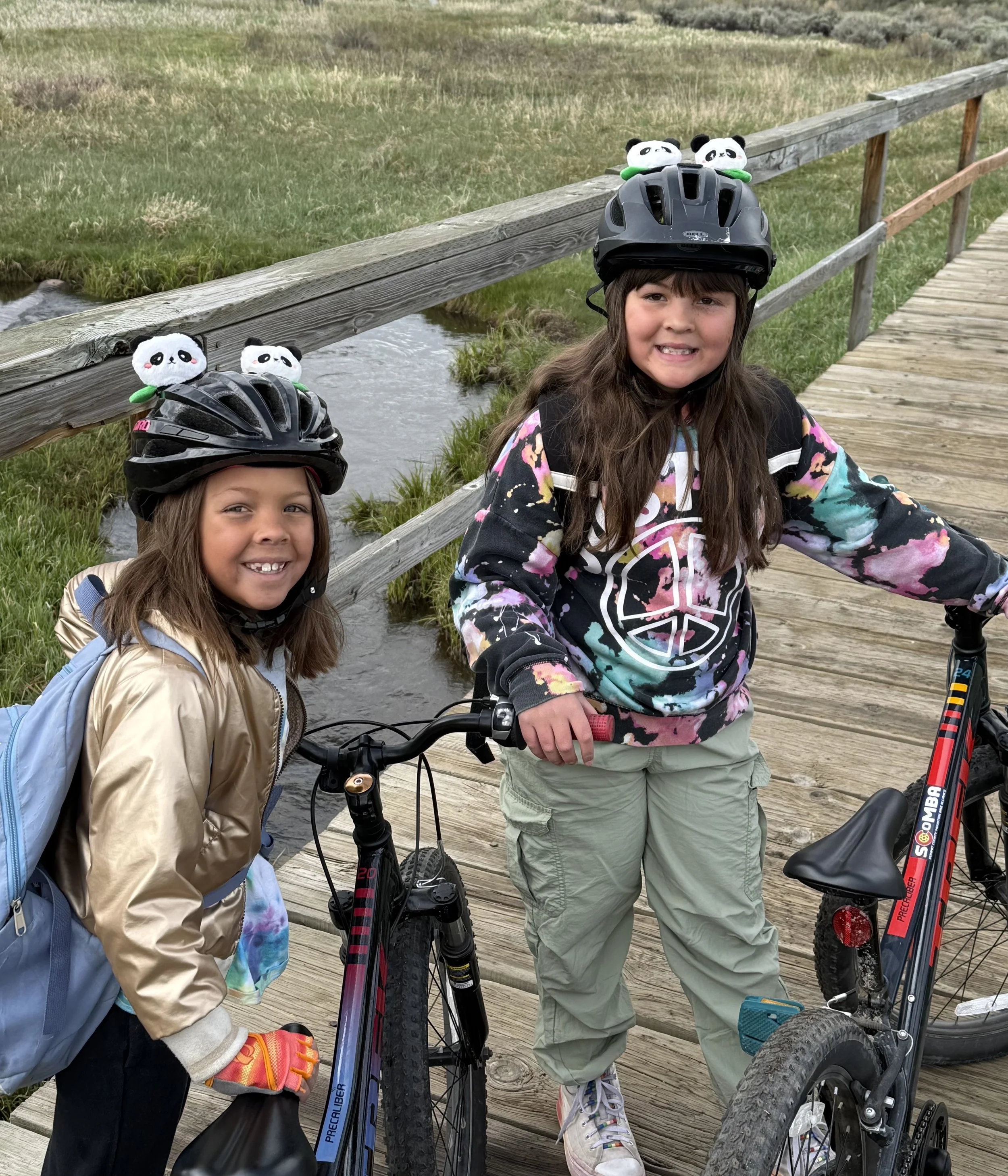 Two young girls with bikes and panda-themed helmets on a wooden bridge over a small creek, smiling at the camera.