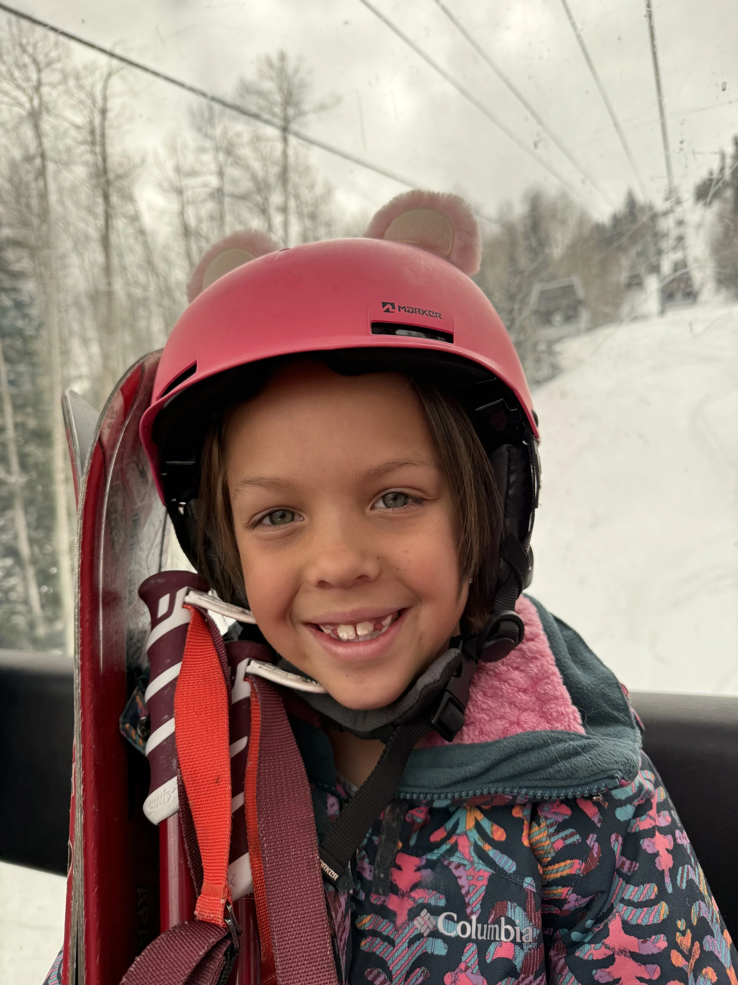 A smiling young girl wearing a pink helmet with bear ears, sitting in a ski lift with snow-covered trees and buildings visible in the background.