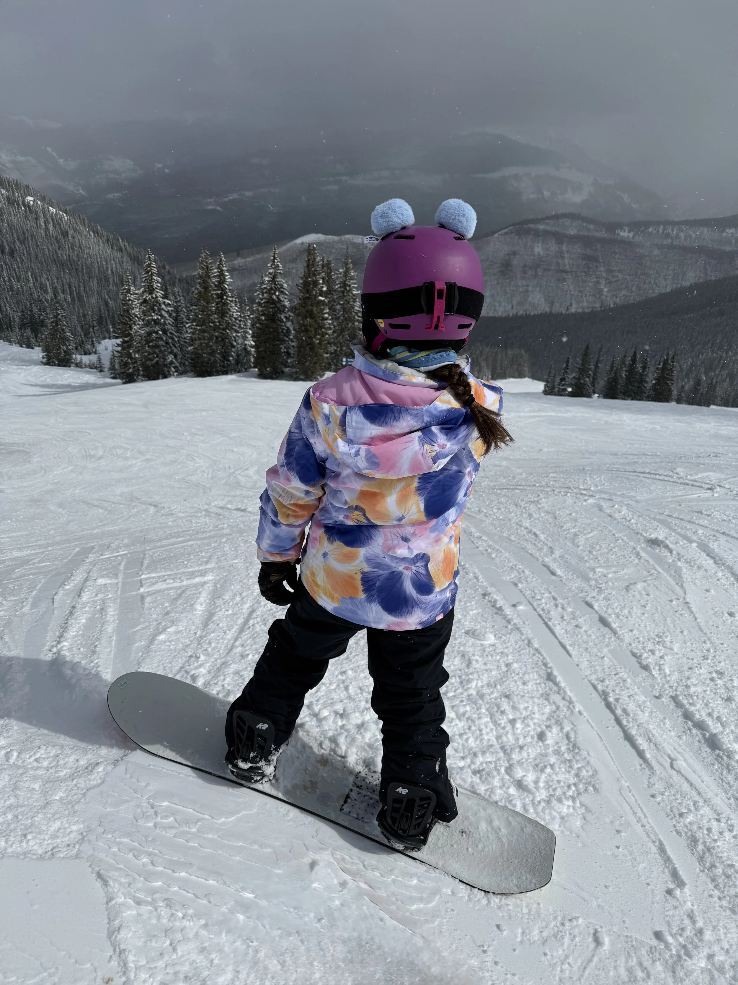 Child in a colorful jacket and black snow pants standing on a snowboard on a snowy mountain slope, facing away, wearing a purple helmet with blue pom-poms, and surrounded by snow-covered trees with cloudy mountain scenery in the background.