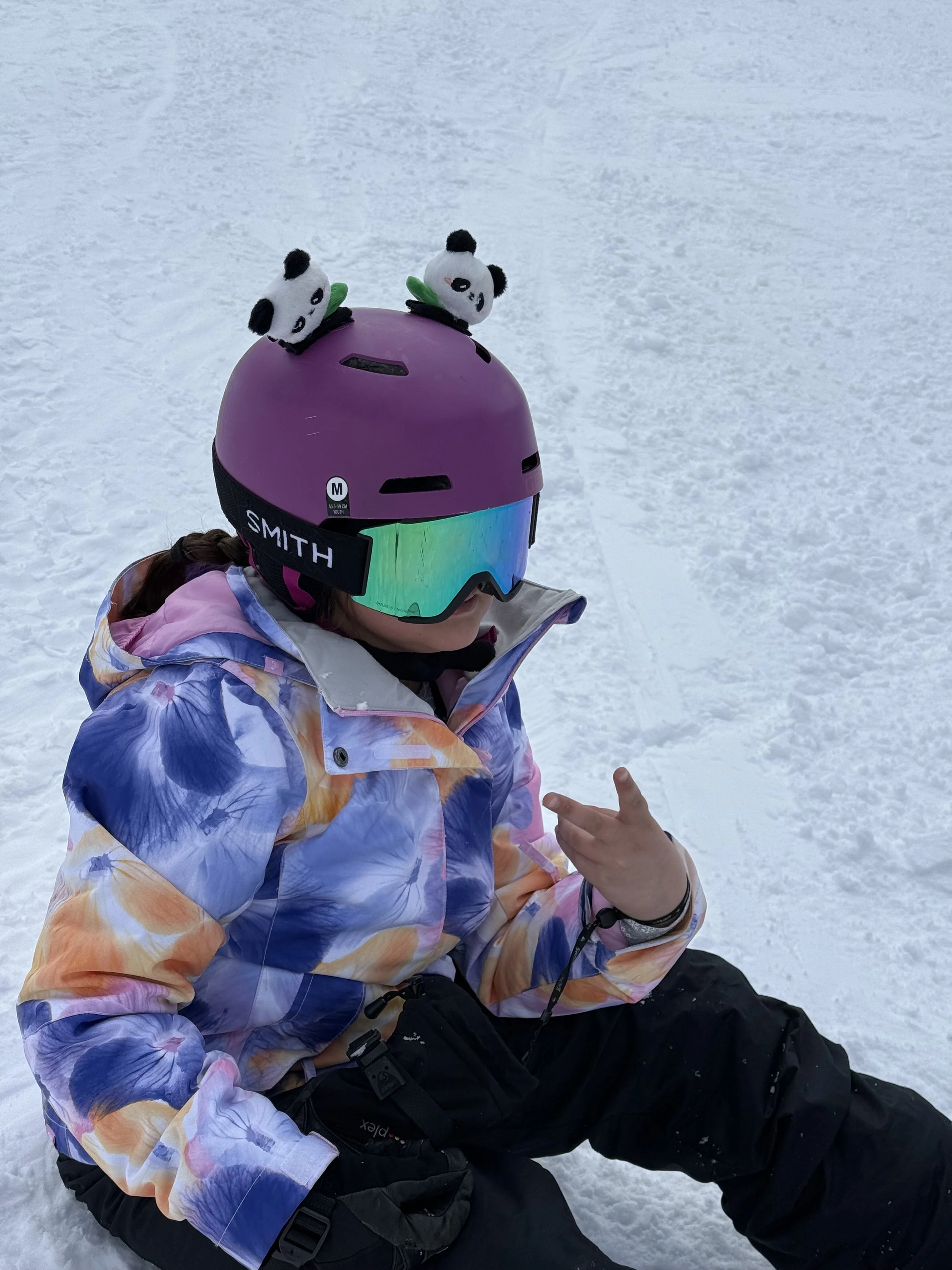 Child in colorful jacket and black snow pants sitting on snow, wearing a purple helmet with panda plush toys on top and reflective ski goggles.