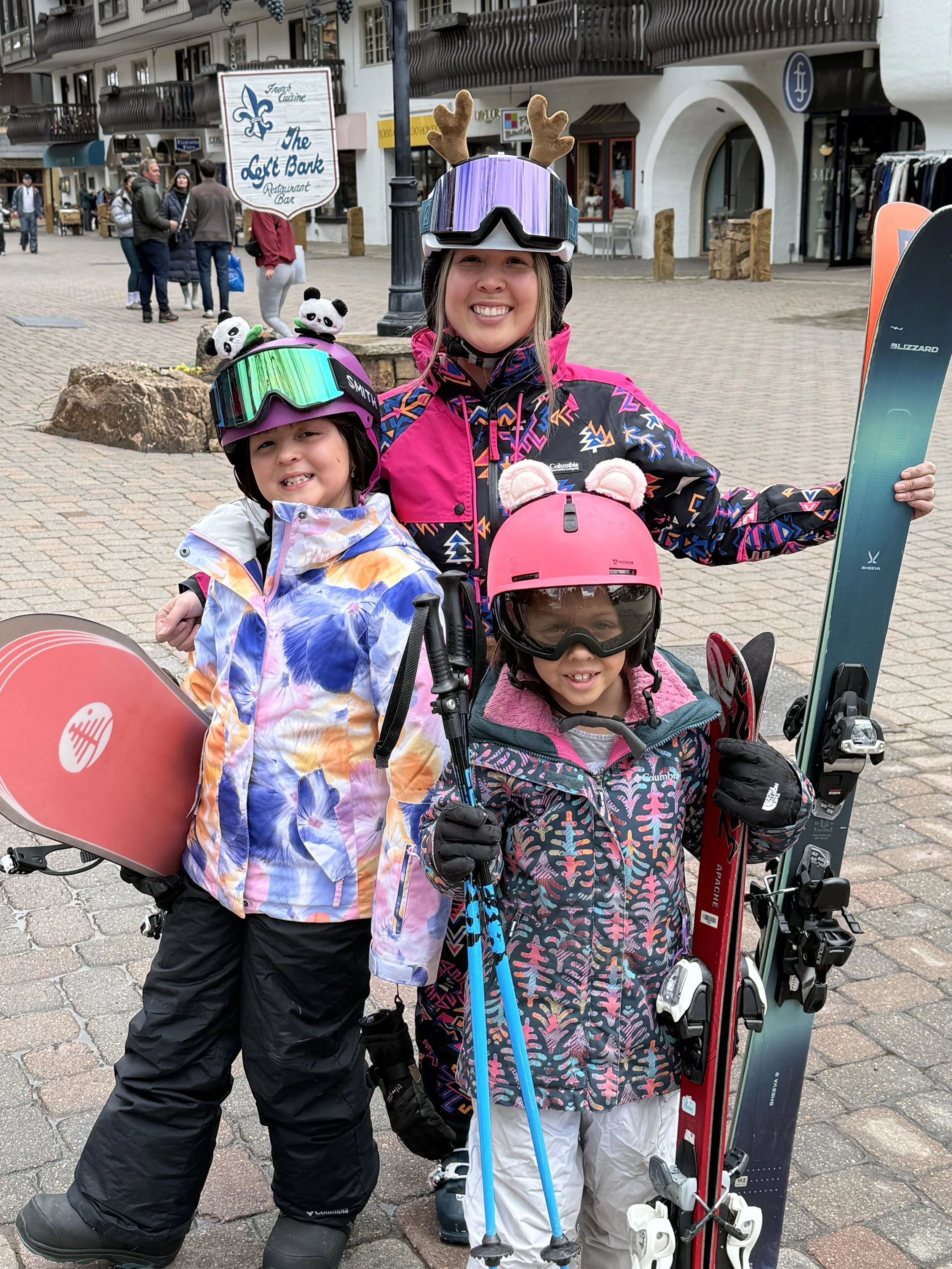 Three children and an adult in ski gear, smiling, standing outdoors with shops and pedestrians in the background. They are holding skis, poles, and a snowboard, and wearing helmets and goggles. The adult has reindeer antlers on the helmet.