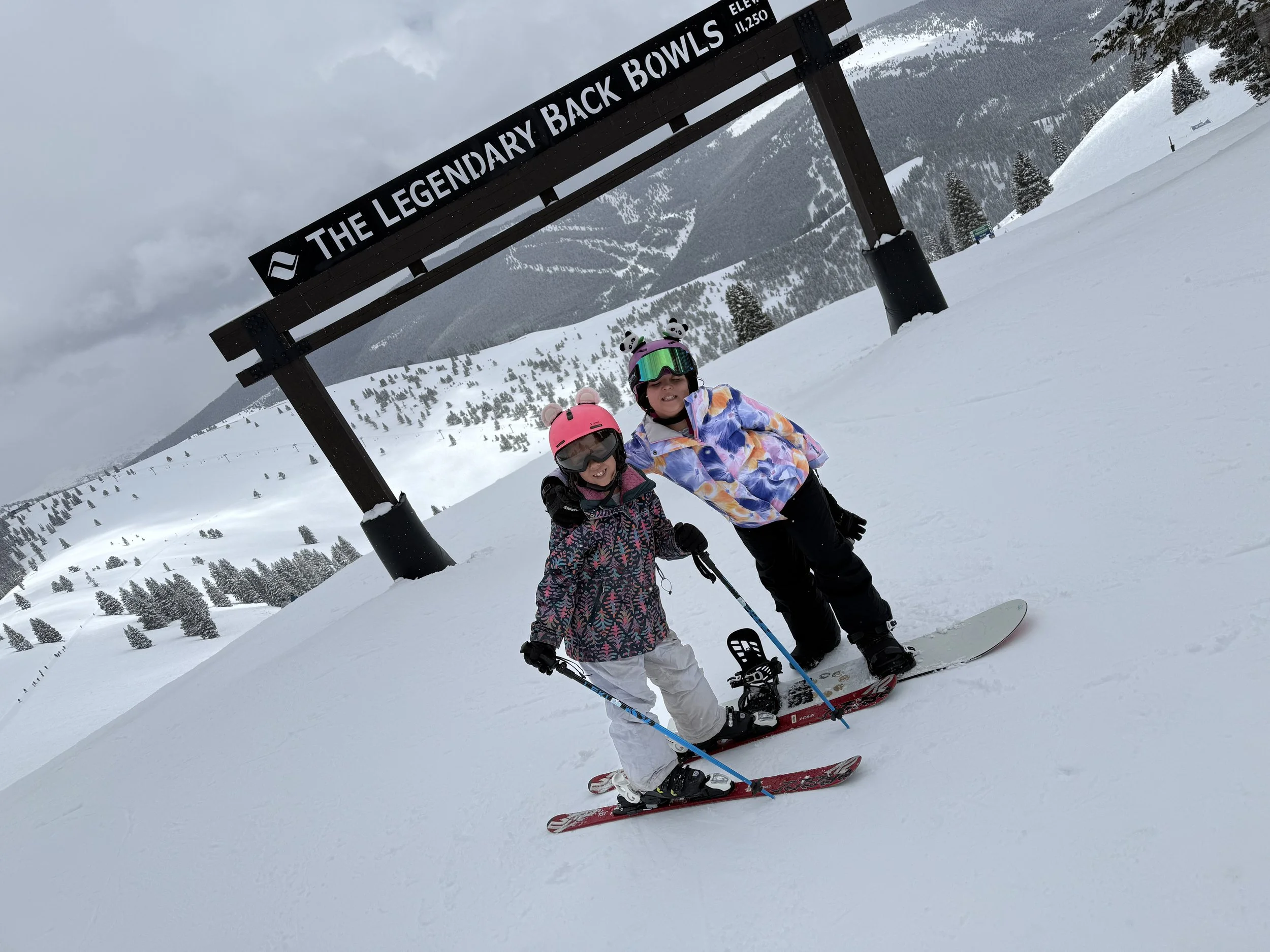 Two children in colorful winter clothing and ski gear standing together on a snowy slope under a sign that reads "The Legendary Back Bowls" with snow-covered mountains and trees in the background.
