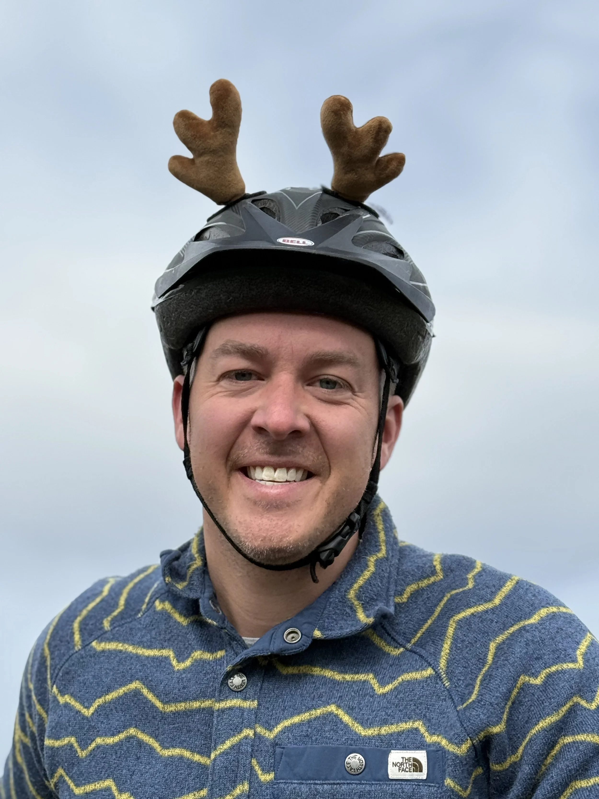A smiling man wearing a bike helmet with reindeer antlers on top, standing outdoors against a cloudy sky.
