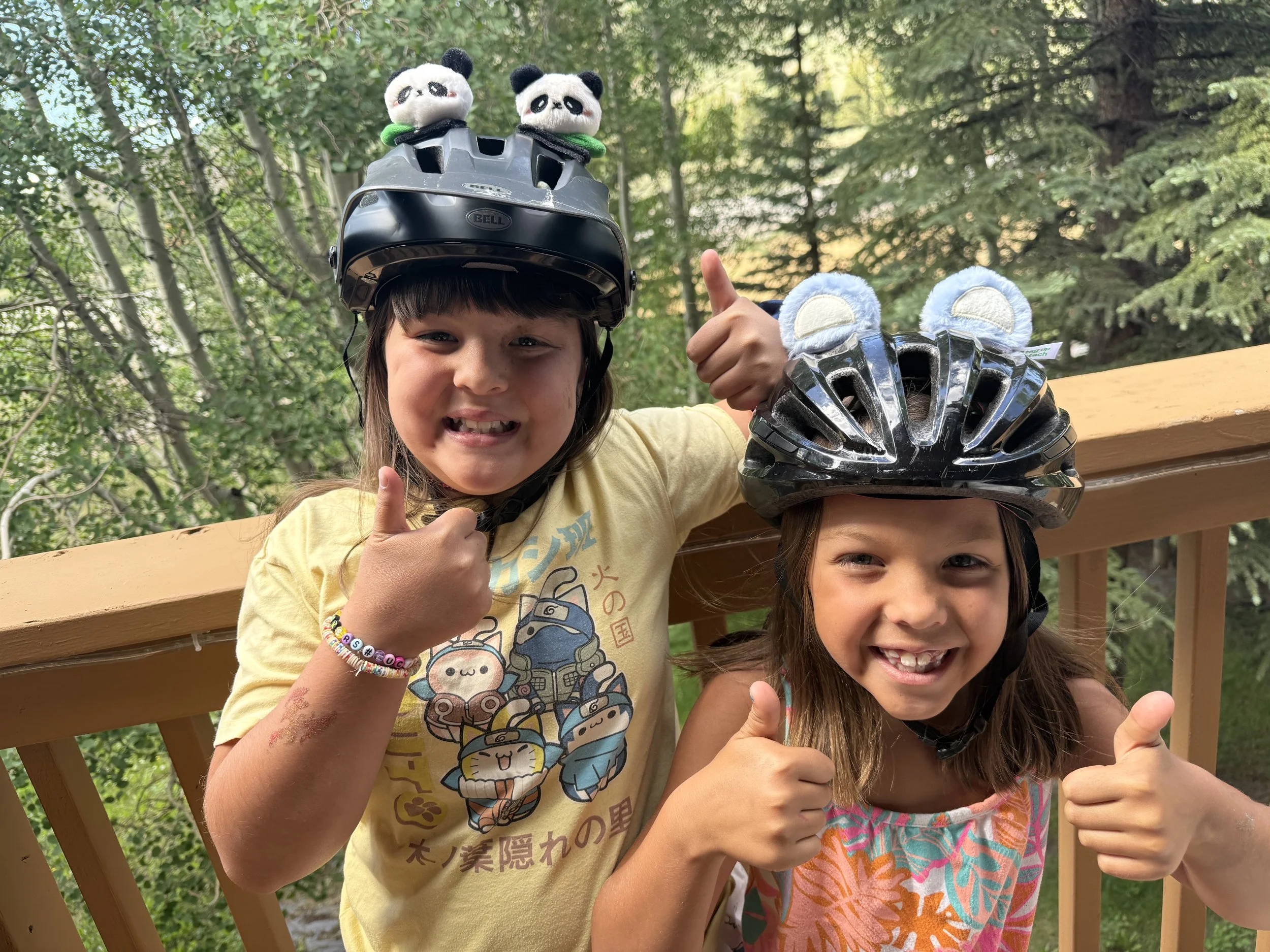 Two young girls wearing bike helmets, giving thumbs up, smiling, outdoors near a wooden railing with trees in the background.