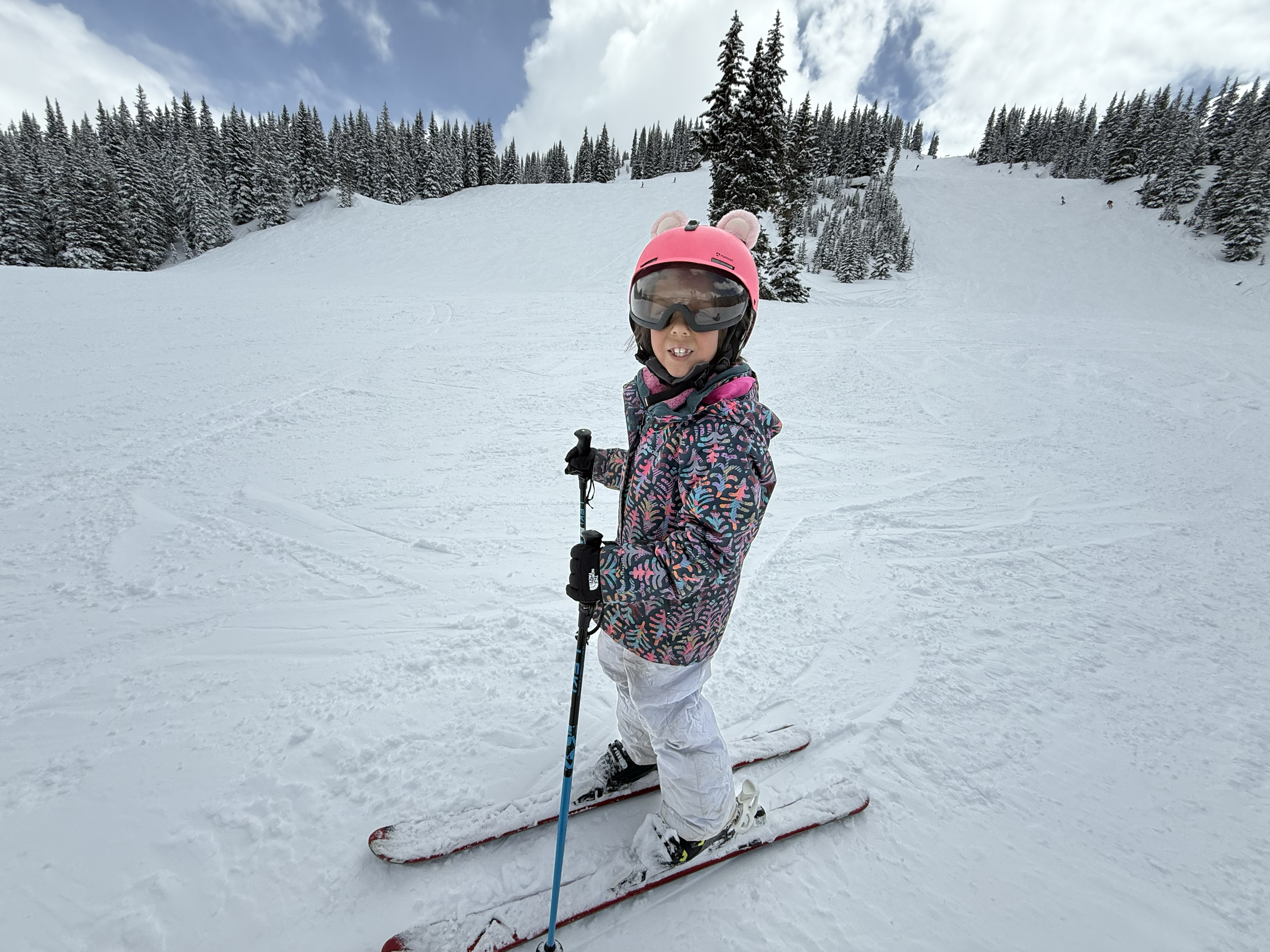 A young girl in a pink helmet with bear ears, sunglasses, a colorful winter jacket, white snow pants, and ski boots standing on skis on a snowy mountain slope with pine trees in the background.
