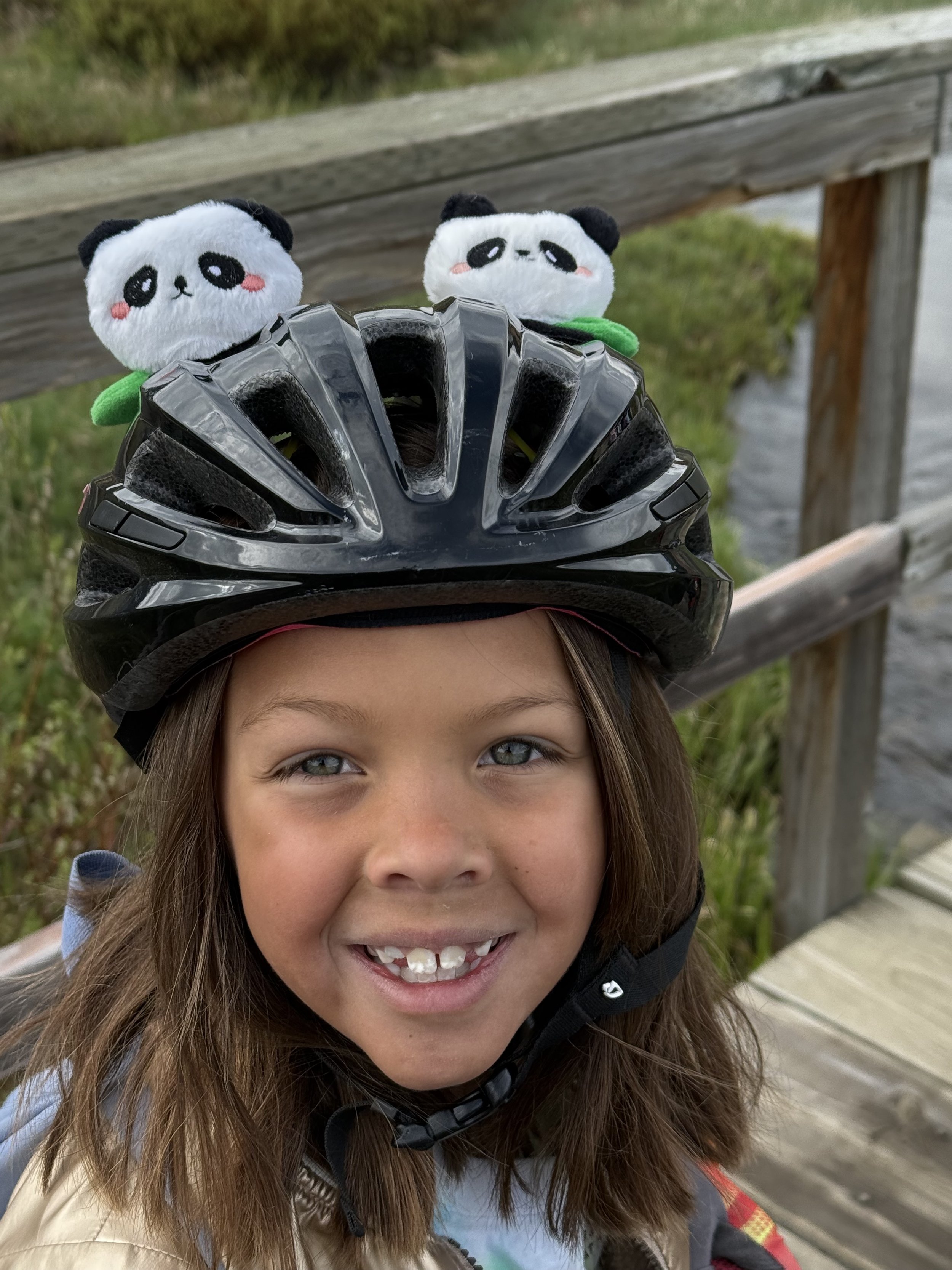 A young girl wearing a bicycle helmet with panda plush toys attached on top, standing on a wooden bridge with water and greenery in the background.