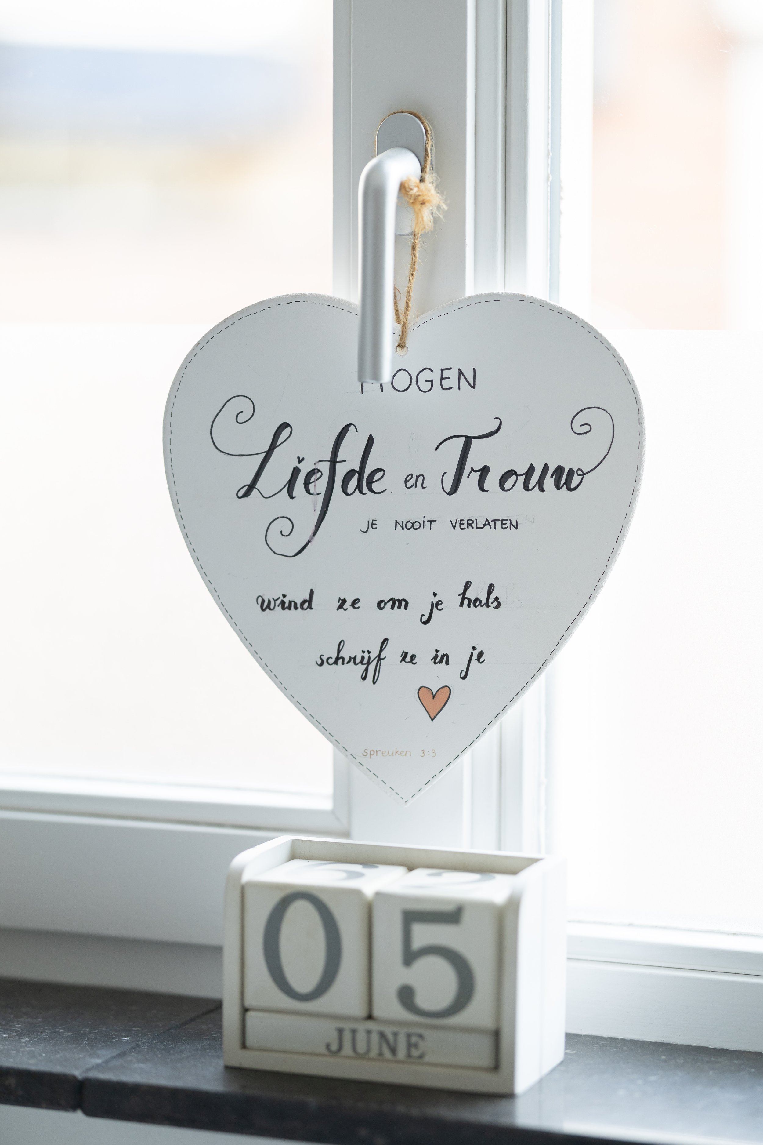 Decorative heart-shaped sign hanging on a window handle with a message in Dutch and a small calendar showing June 5th, placed on a window sill.
