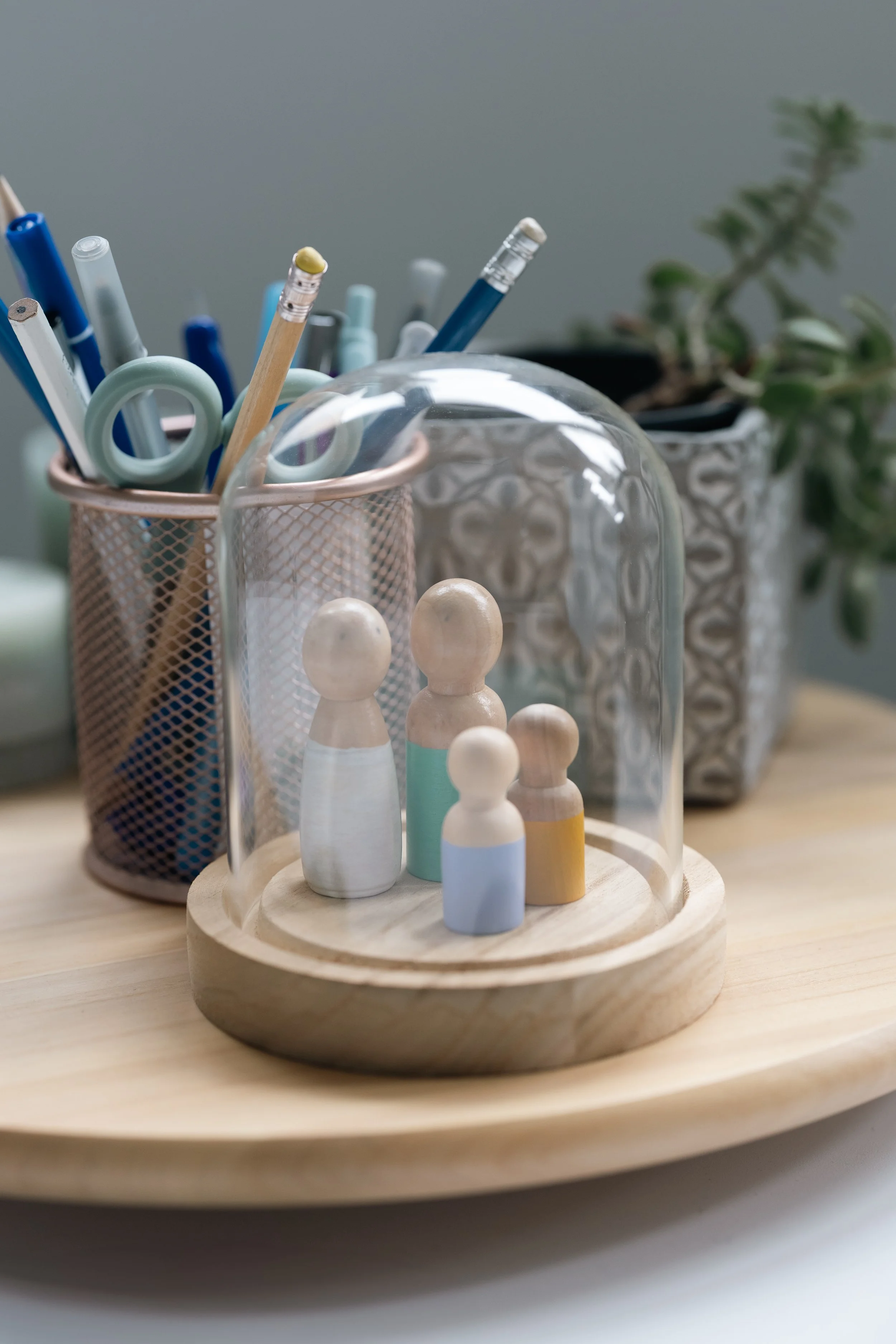 Decorative wooden figurines under a glass dome on a wooden tray, with stationery items and a potted plant on a desk.