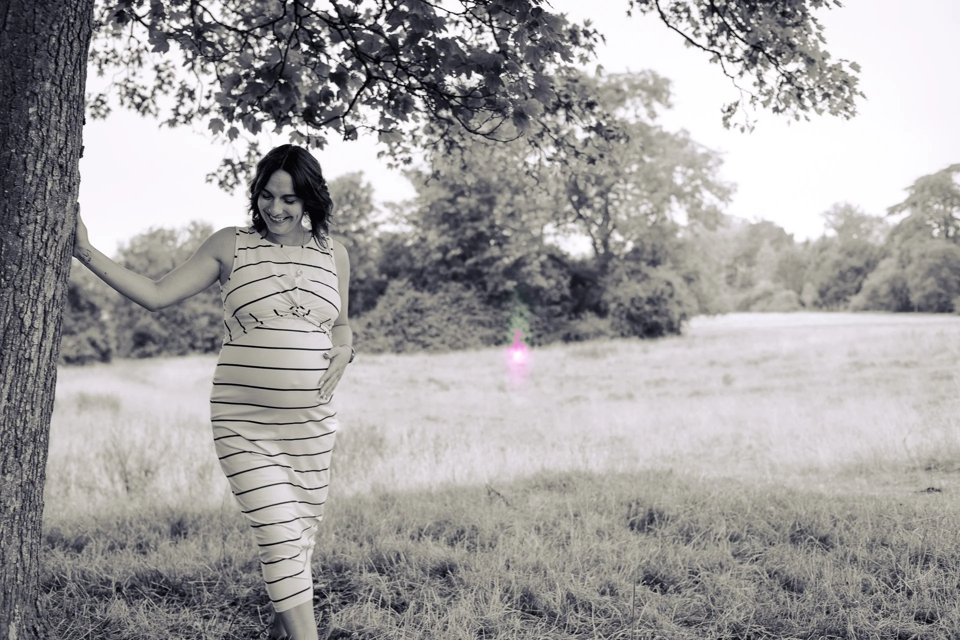 A woman in a striped dress standing outdoors near a tree, smiling, with a grassy field and trees in the background.