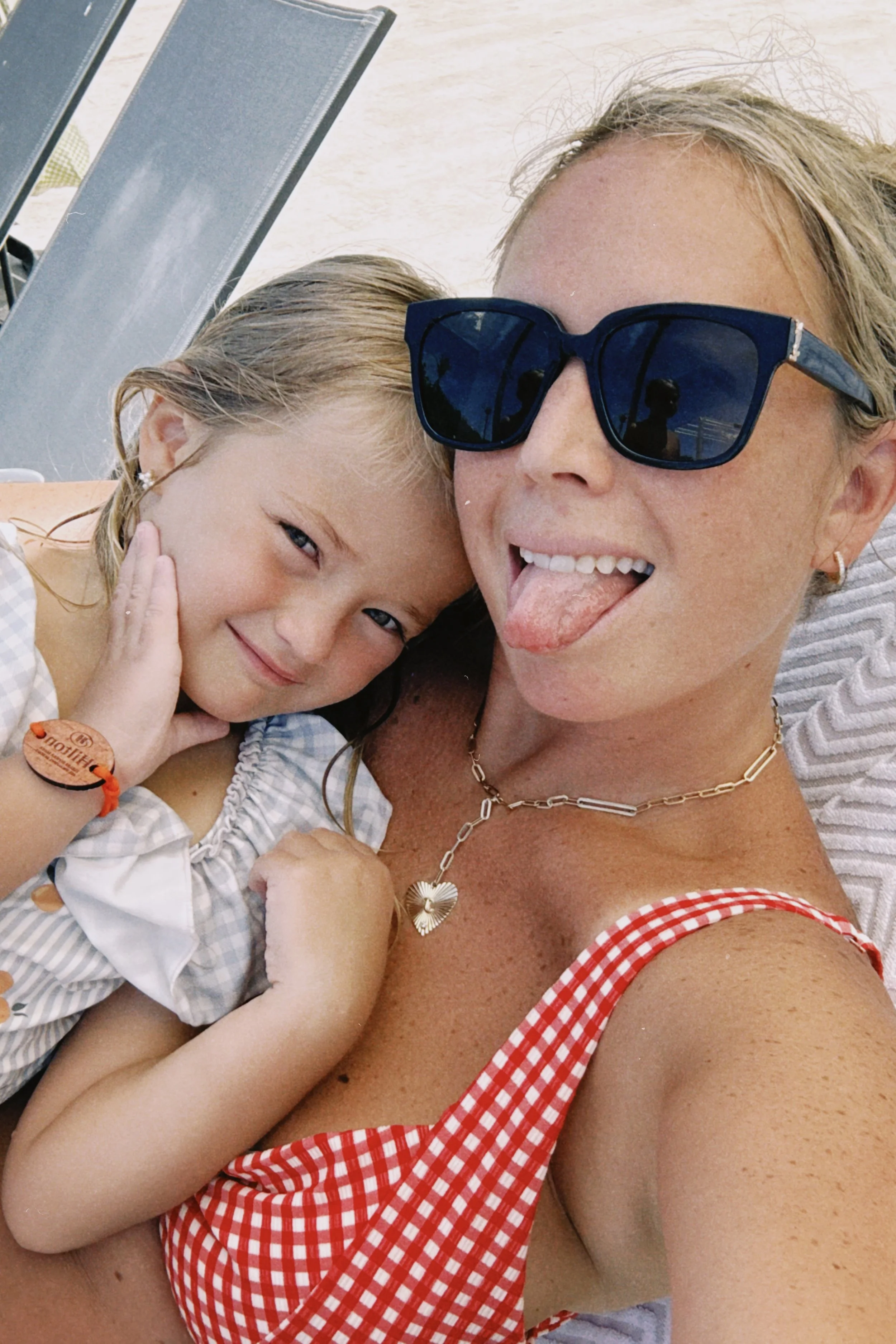A woman and a young girl taking a selfie at the beach. The woman is wearing sunglasses and a red and white checkered swimsuit. The girl has a big smile and is close to the woman, who is sticking out her tongue.