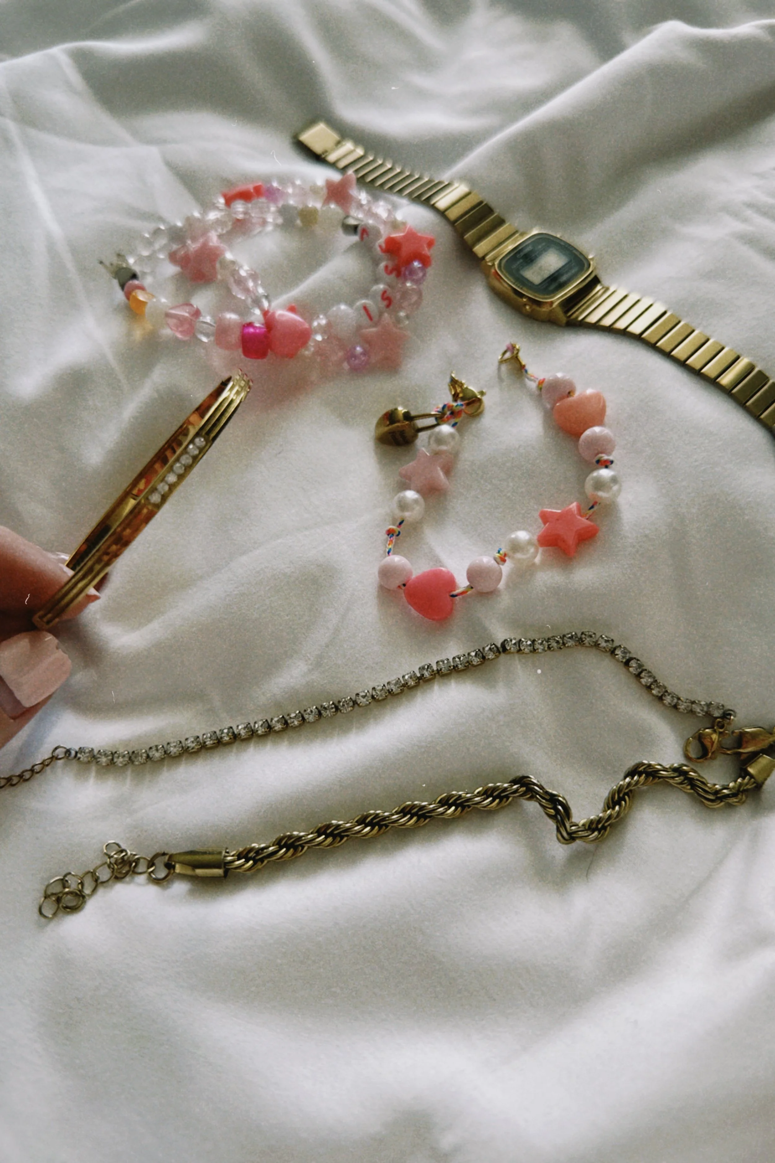 Collection of jewelry including a gold watch, pink and white beads bracelet, pearl and star necklace, and gold and silver chains on a white fabric surface.