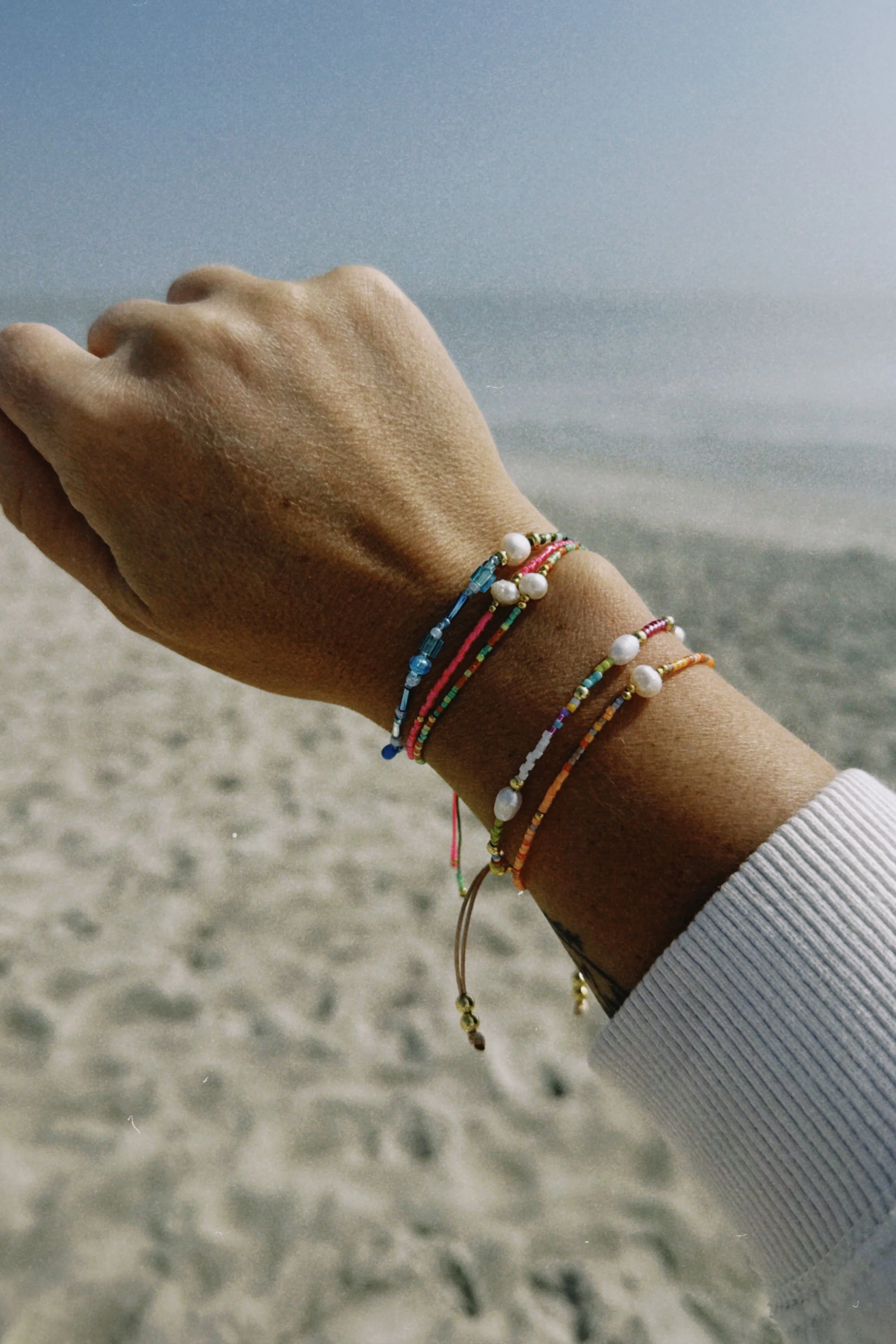 Close-up of a person's wrist with colorful beaded bracelets and a white pearl bracelet, against a beach background with sand and the ocean.