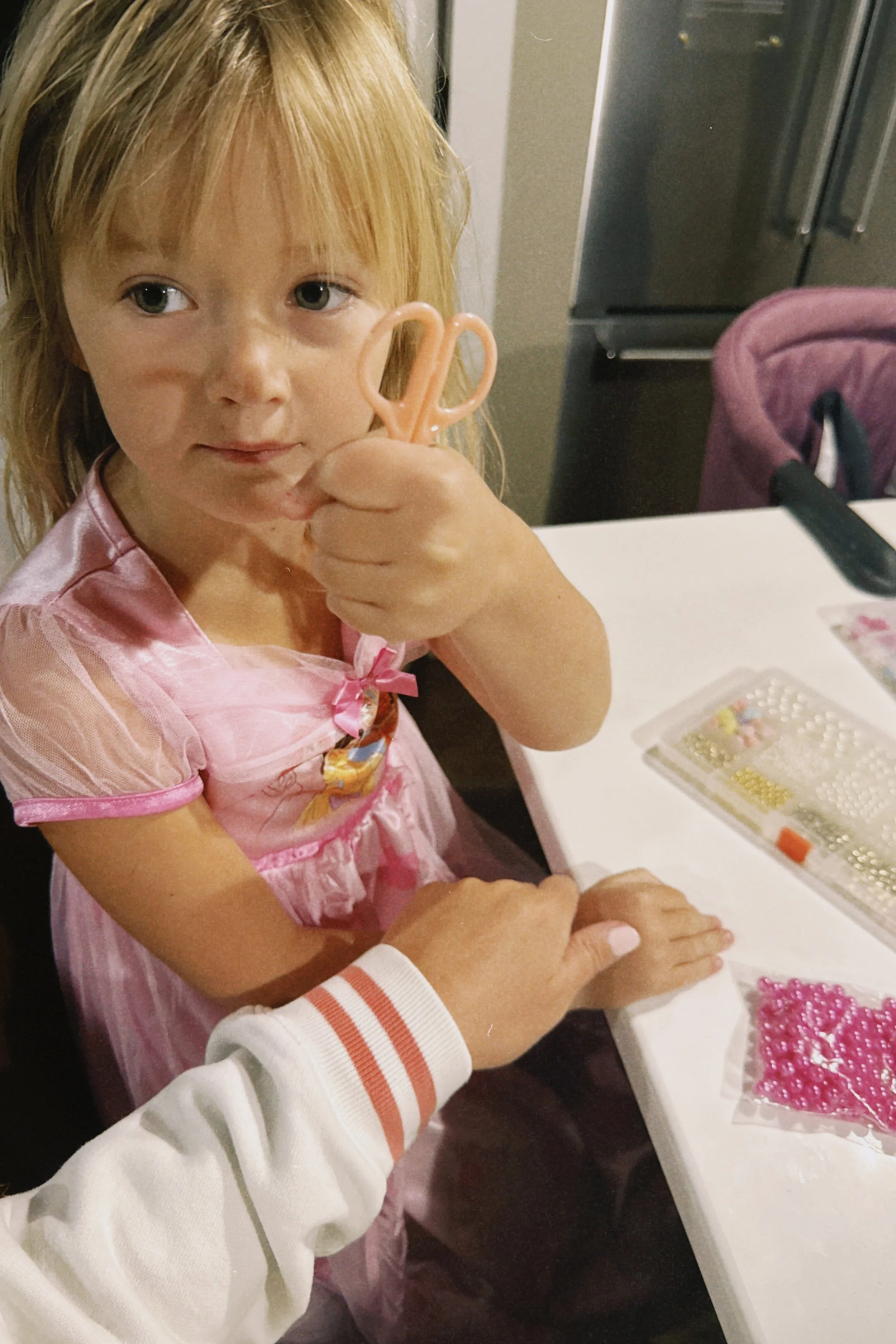 A young girl with blonde hair and green eyes, wearing a pink dress with a character print, is holding a small pair of scissors near her face and smiling slightly. She is seated at a table with craft supplies, including beads and tools, and an adult's hand is resting on her arm.