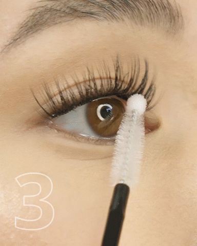 Close-up of a person applying mascara to their upper eyelashes with a white spoolie brush.