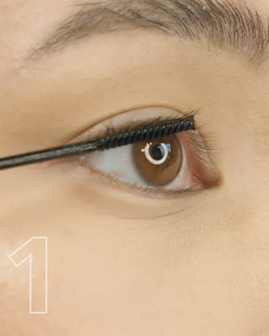 Close-up of a person applying mascara to their long eyelashes in front of a mirror.