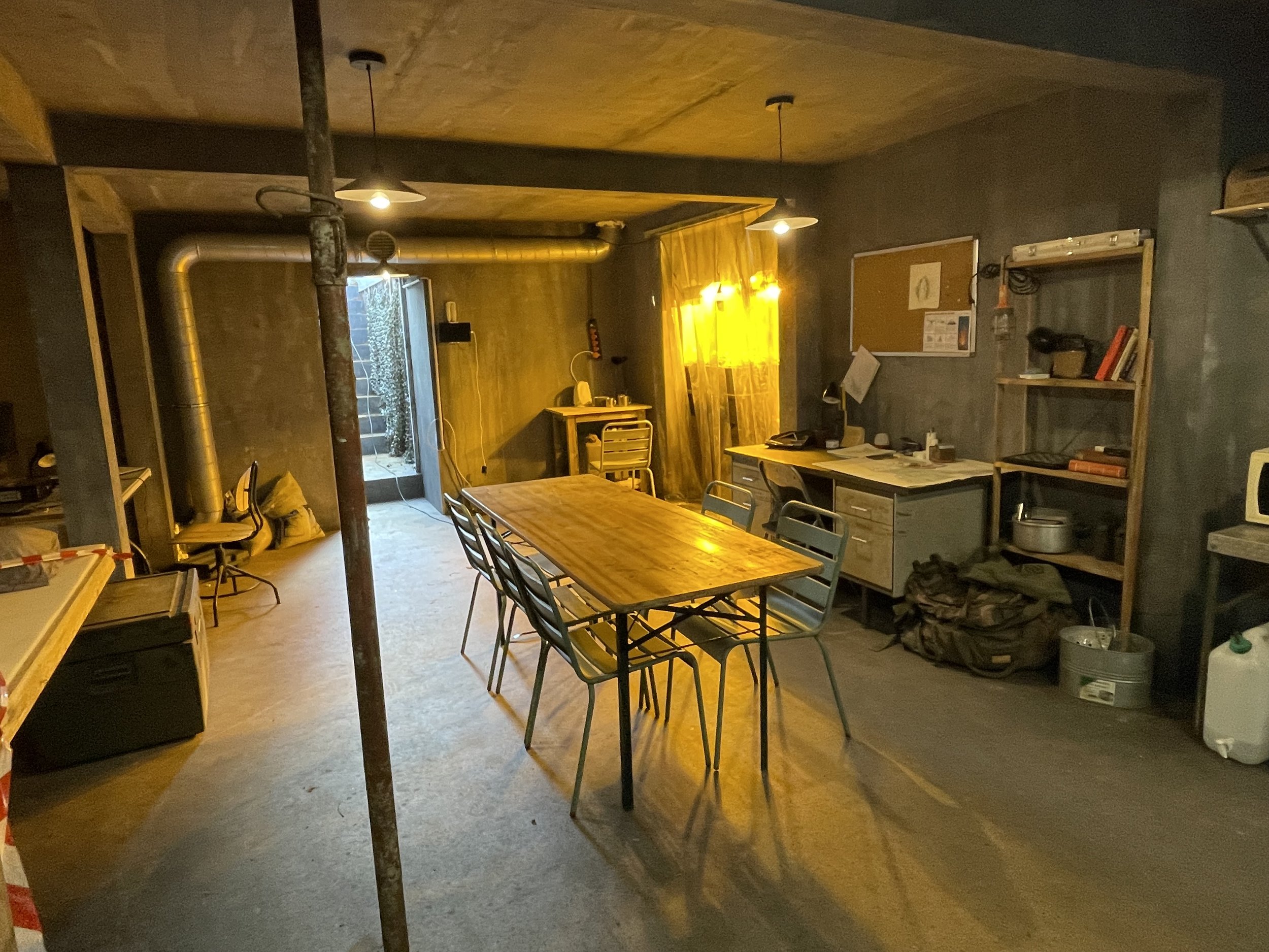 Interior of a rustic, industrial-style room with a wooden table, blue chairs, a desk with papers, and shelves with books and kitchen items. An open door leads outside, and warm lighting illuminates the space.