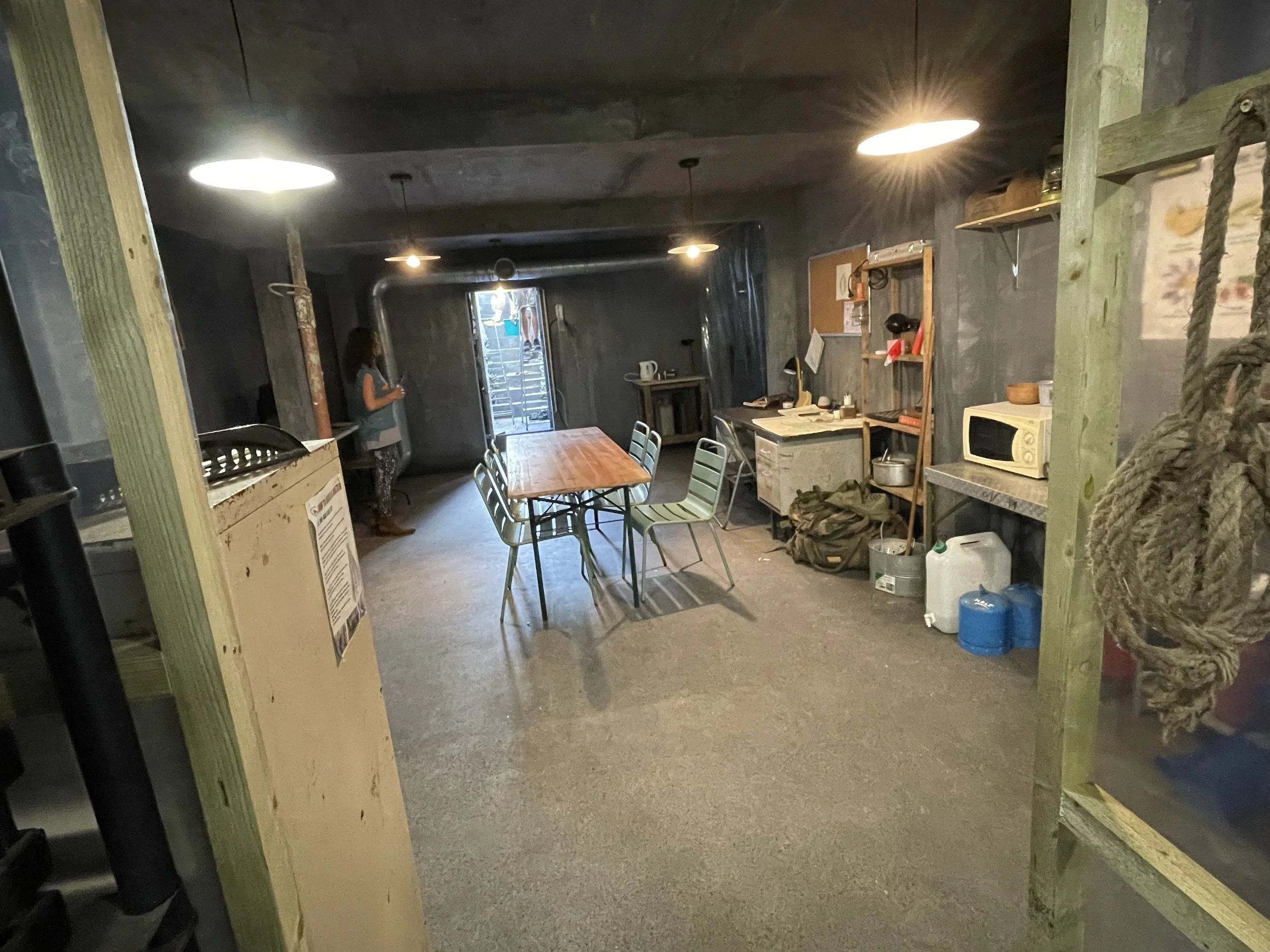 Interior of a rustic kitchen with a wooden table and green chairs, shelves with kitchen items, and a woman standing near the door, dimly lit with exposed pipes and dark walls.