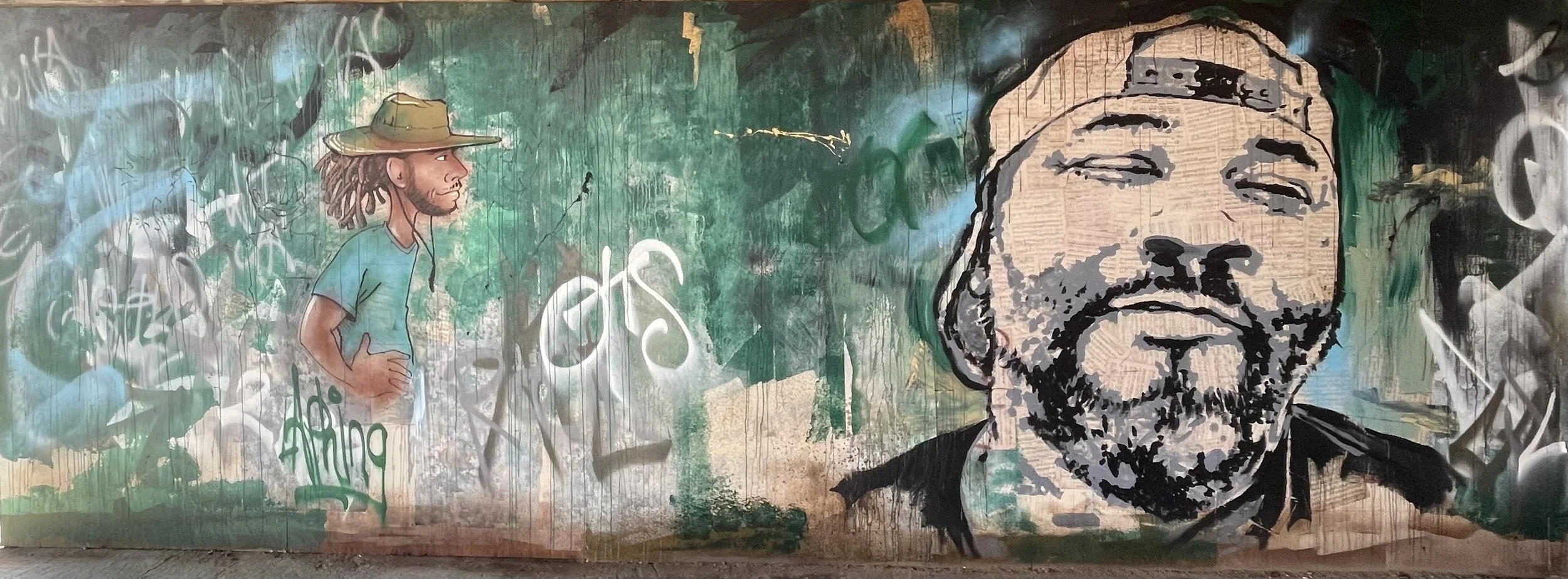 Street art mural of a man with a beard and glasses wearing a cap, smiling, with a background of green and black spray paint. The mural also features a profile of another man with dreadlocks and a hat, and various graffiti tags.