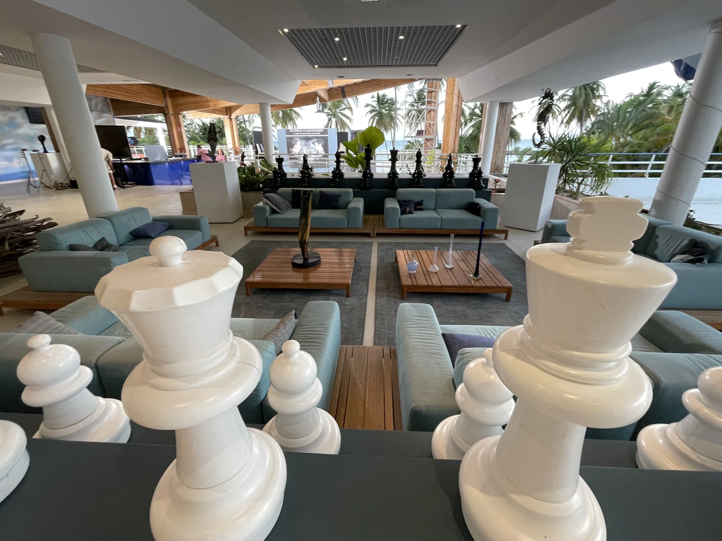 Indoor lounge area with pastel blue sofas, wooden tables, and a large chess set in the foreground, overlooking a waterfront with palm trees.