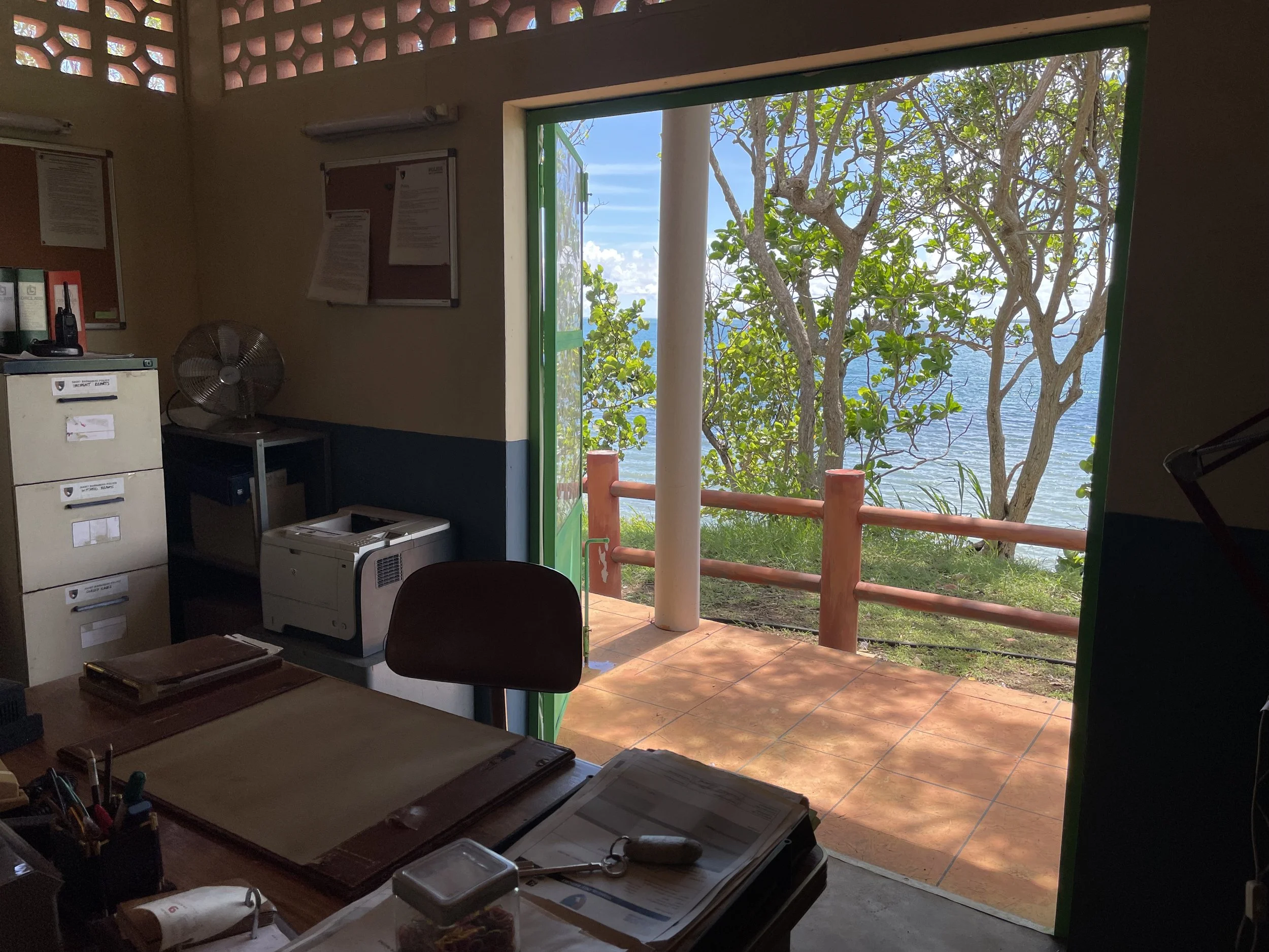 Office with desk and papers, open door leading to outdoor view of trees, ocean, and blue sky