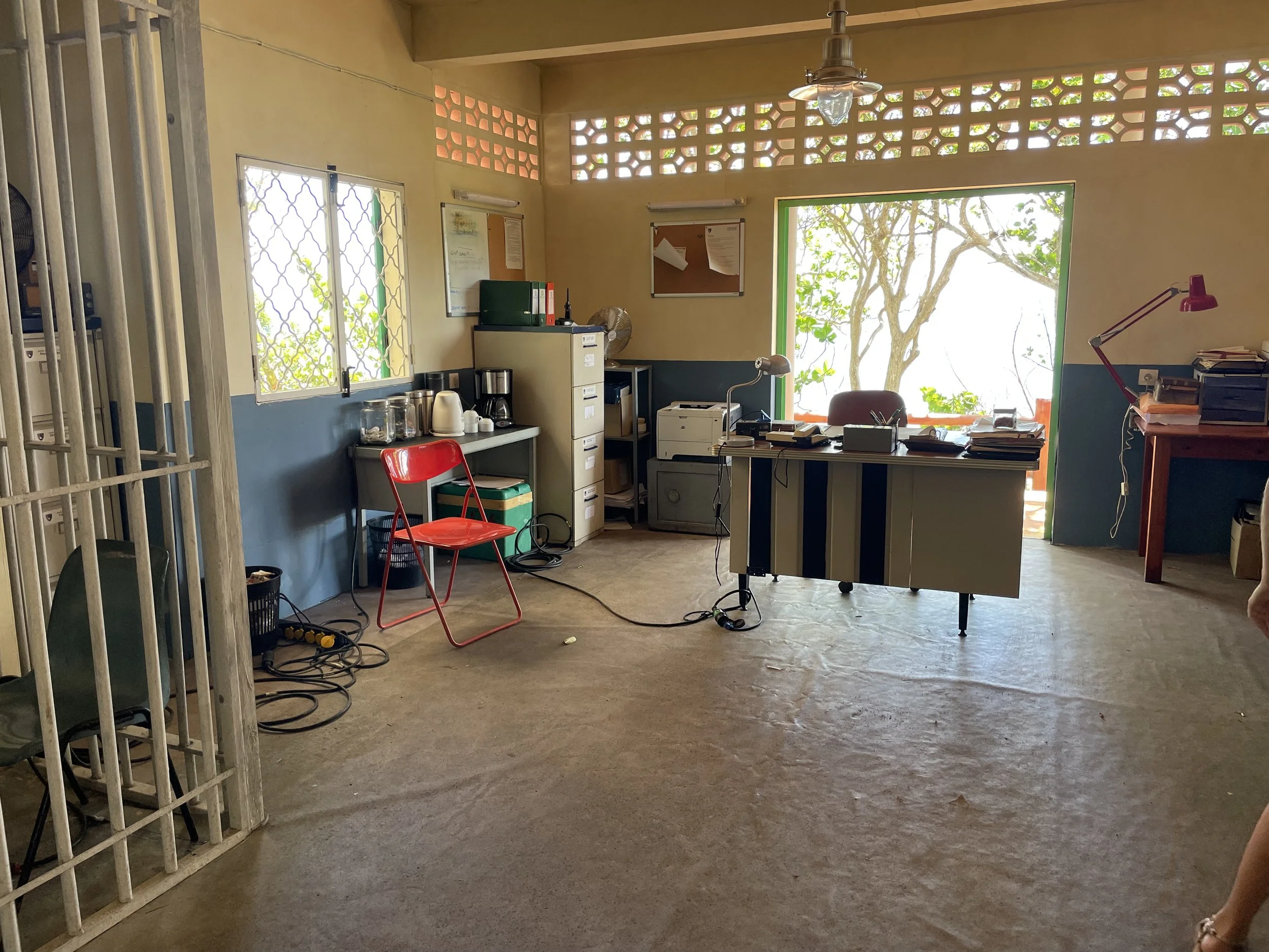 An office with a large window, a desk with stacks of papers, a red desk lamp, a filing cabinet, a small coffee station, and a metal cage on the left side.
