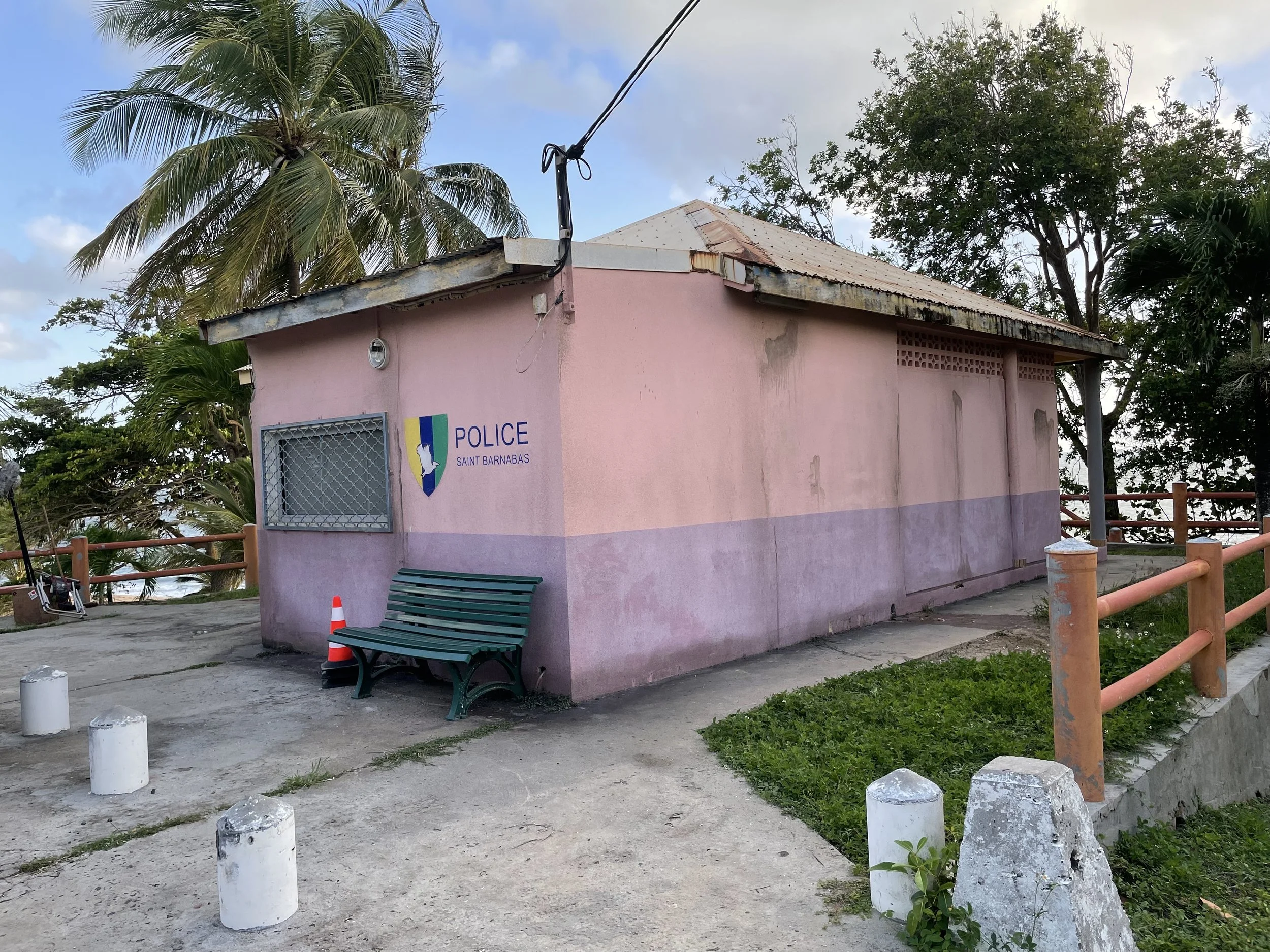 Pink police station building with a sign reading 'POLICE Saint Barnabas,' a green bench outside, surrounded by palm trees and greenery, with a cloudy sky overhead.