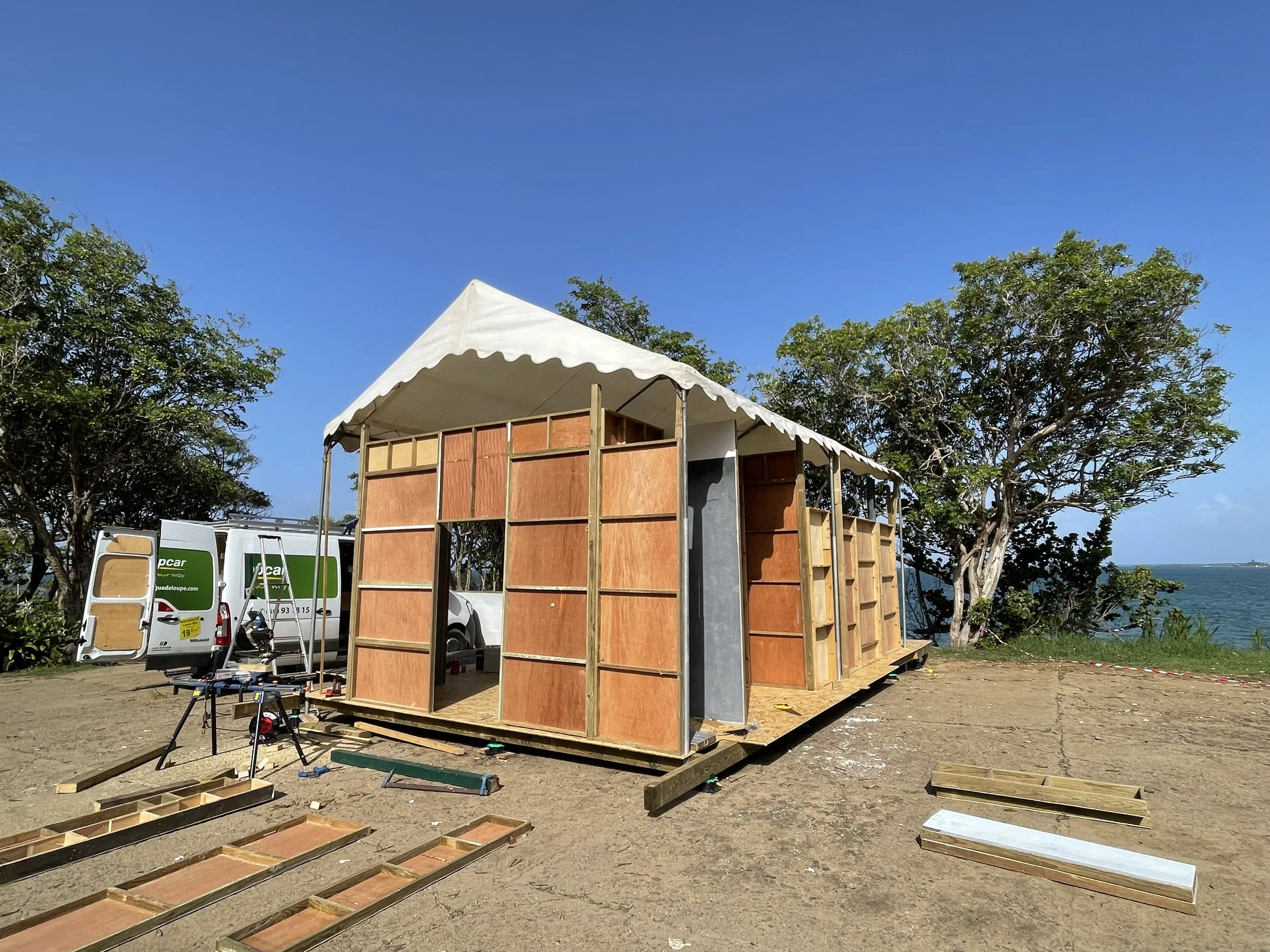 Under construction tiny house with wooden frame and white canopy, on sandy ground near trees and water, construction tools and materials scattered around.