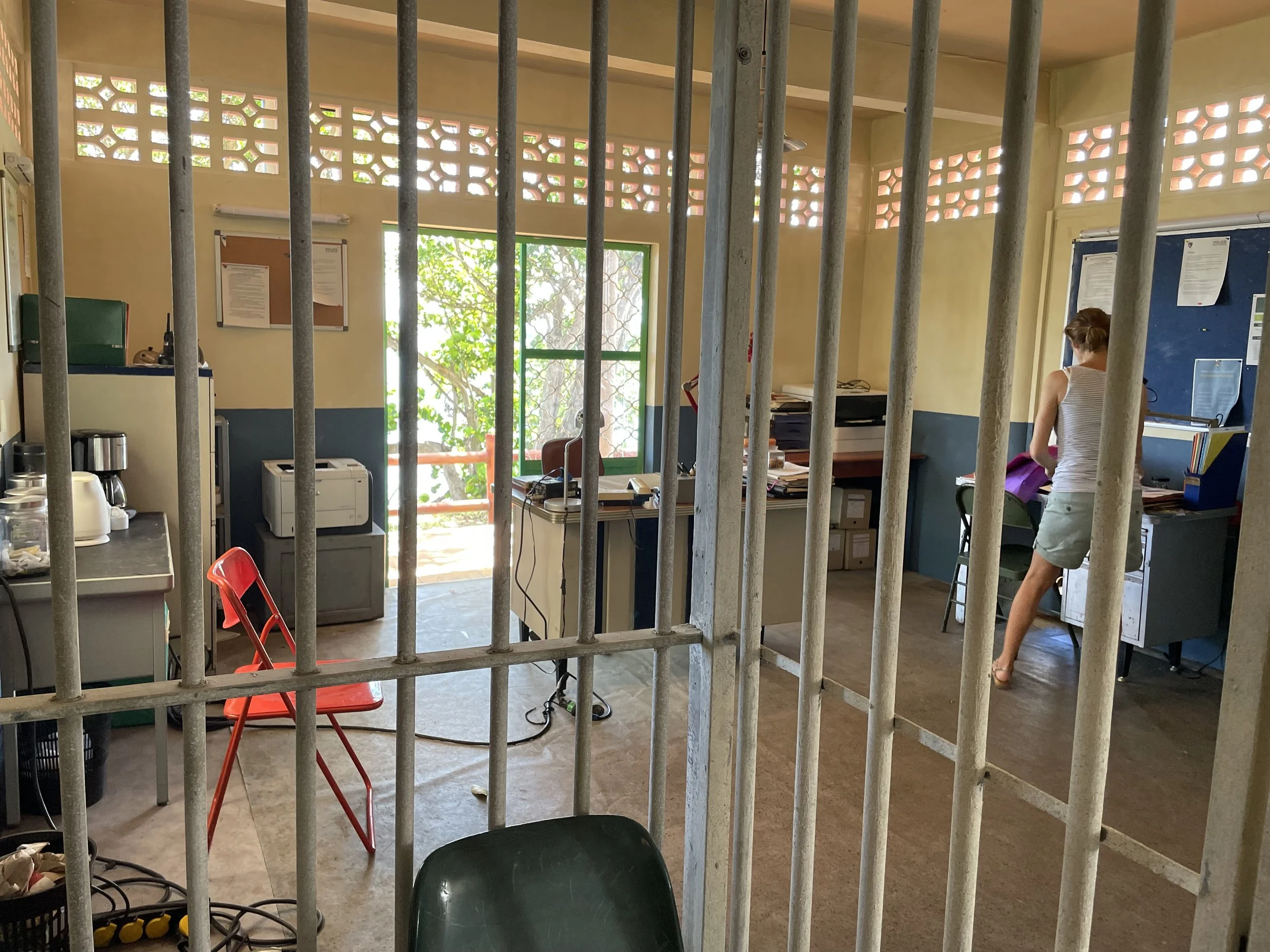 View of an office room behind jail bars, with a woman working at a desk, a chair, a red stool, and various office supplies and equipment.