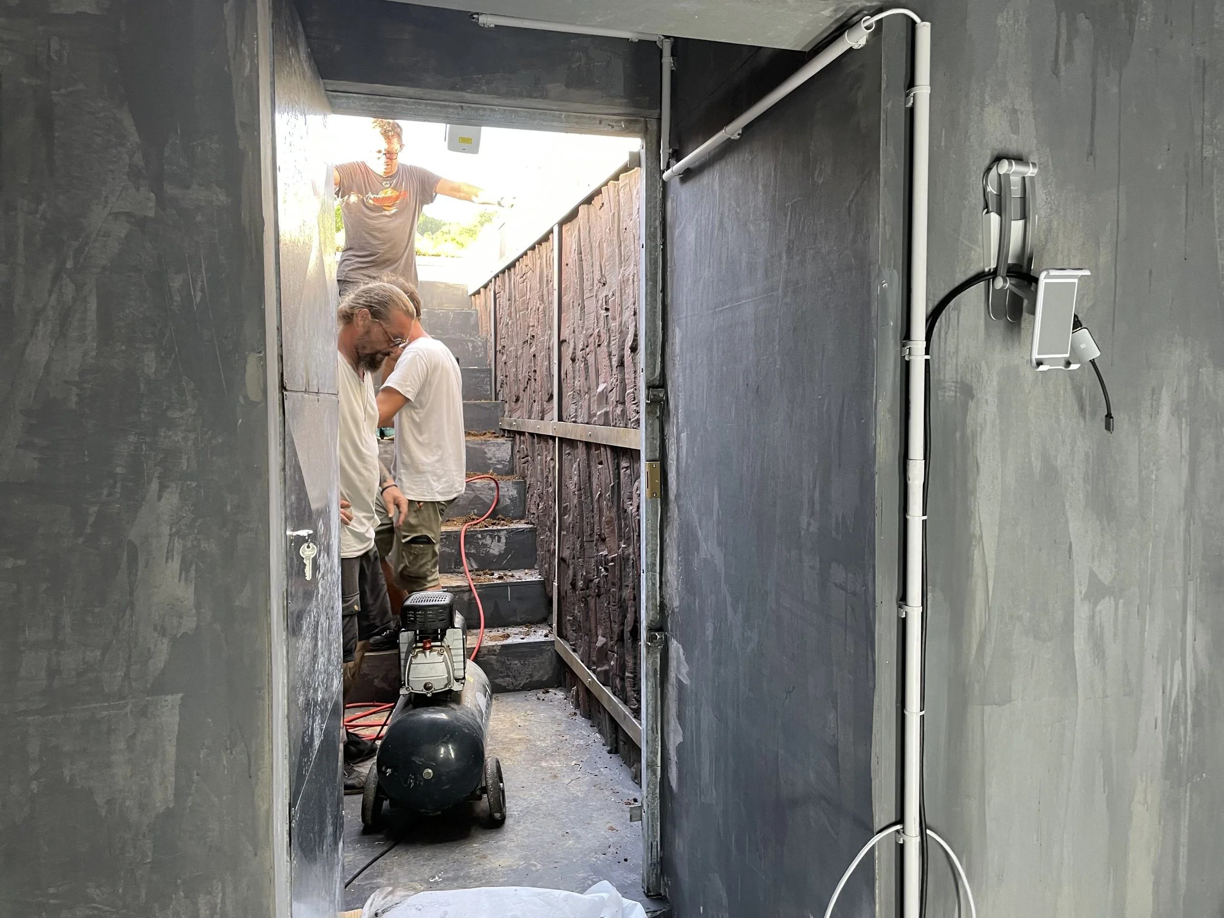 Three men working outside a modern house, with one man inside near the entrance, another at the top of stairs, and the third standing near the door. Equipment and tools are present, indicating construction or renovation work.