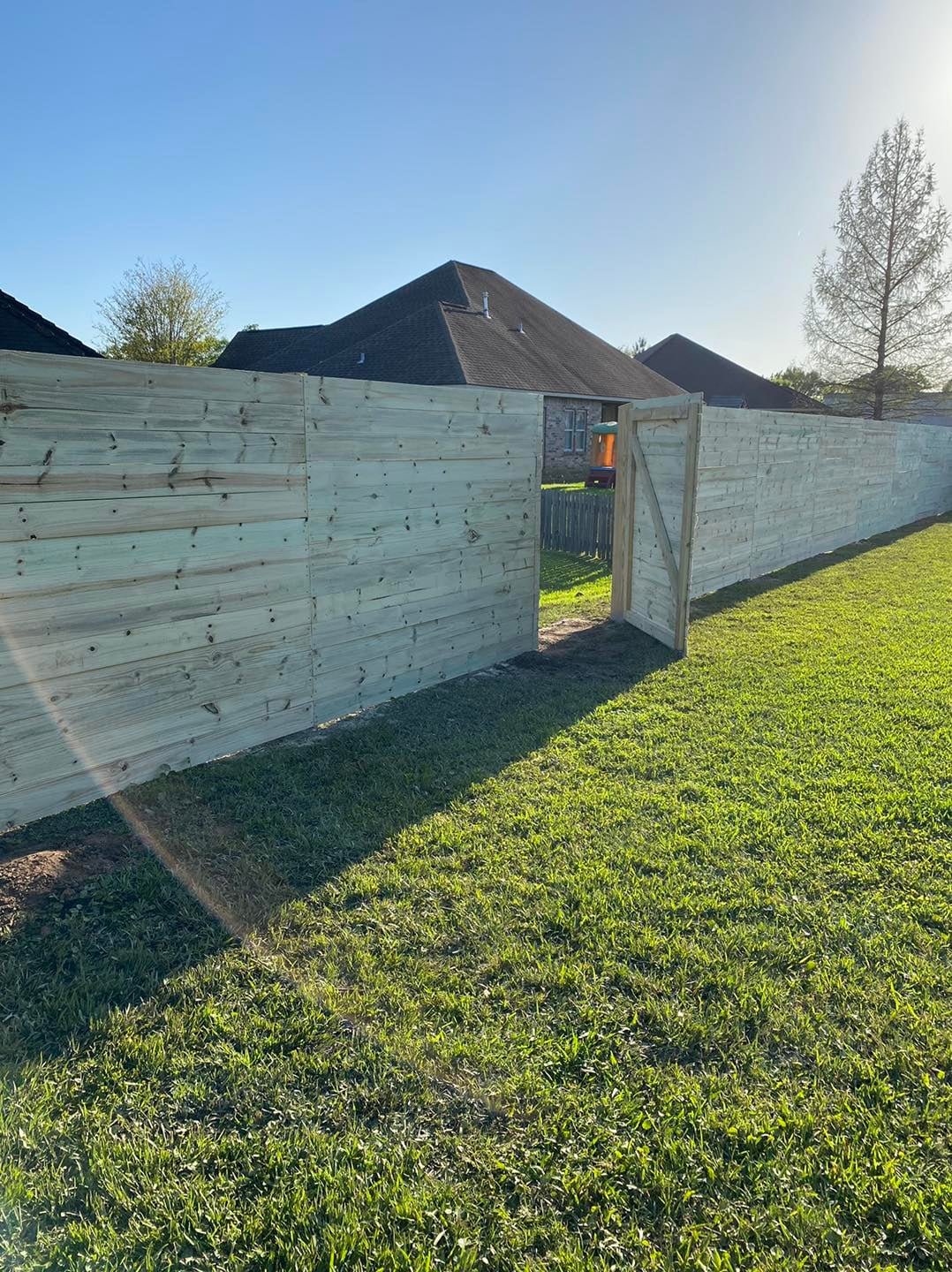 Newly built wooden fence with a gate surrounding a backyard, with houses and trees visible in the background and a clear blue sky.