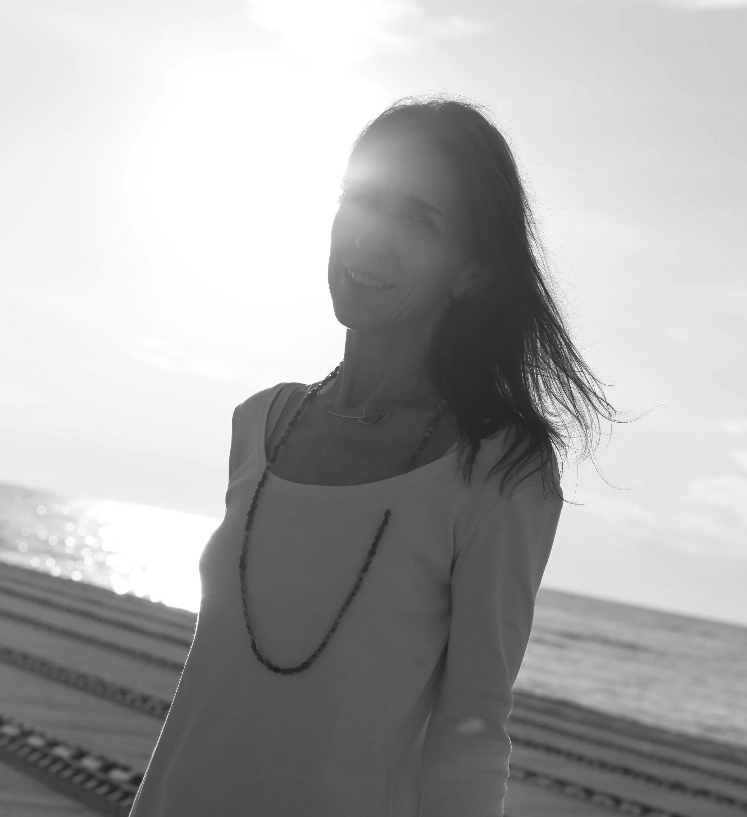 Mujer sonriente de perfil, con cabello largo, usando collar de cuentas, en la orilla del mar con líneas de ferrocarril en el fondo, en un día soleado.