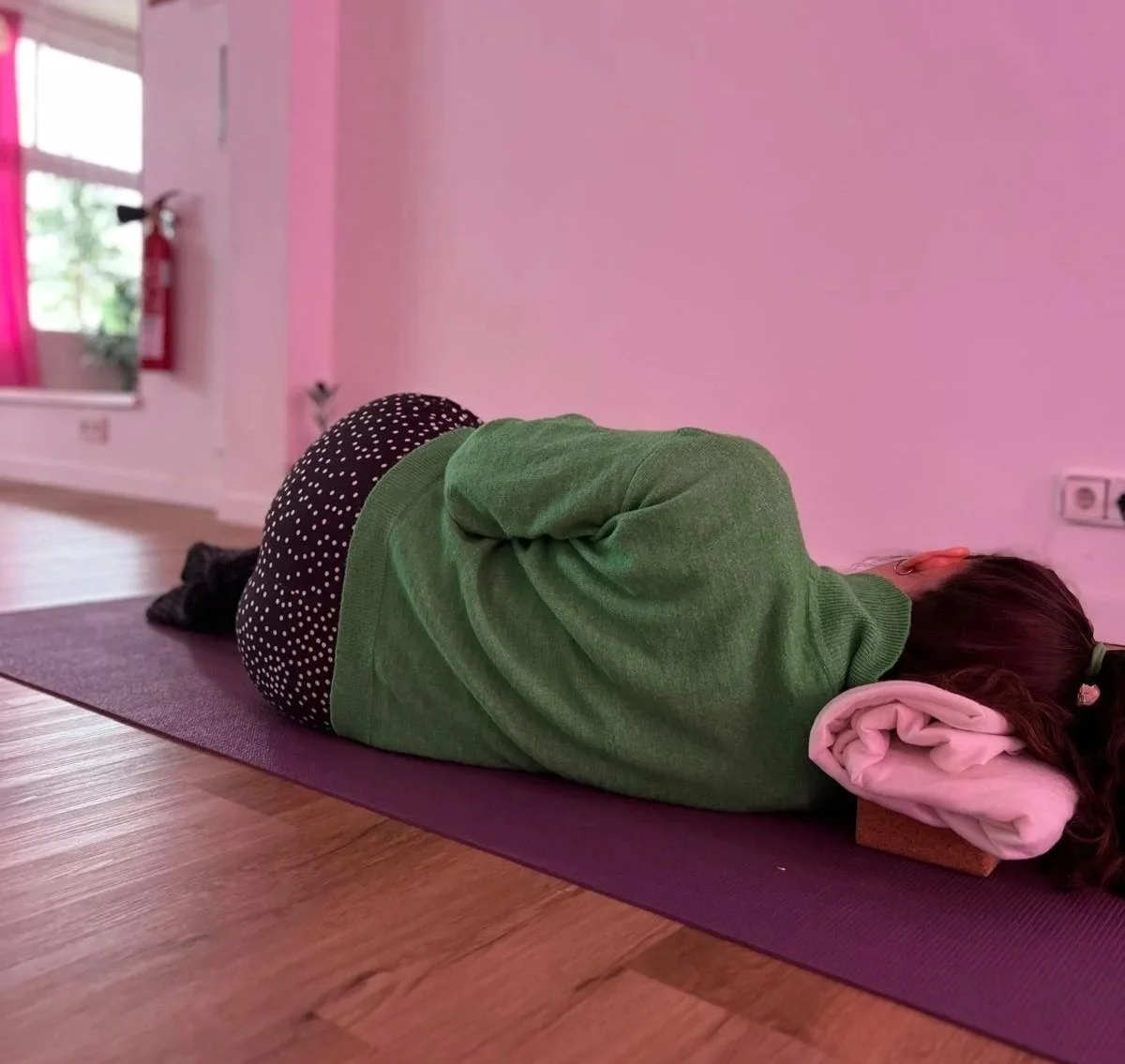 Una niña acostada en una colchoneta de yoga en postura de descanso, con la cabeza sobre un rodillo de espuma, en una habitación con paredes rosas y piso de madera.