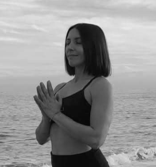 Una mujer con cabello corto, en posición de oración, frente al mar en un día nublado.