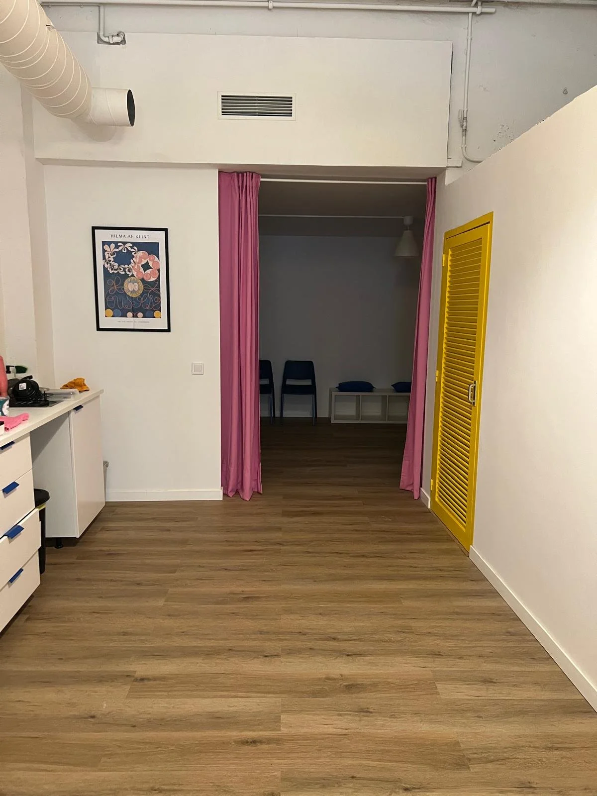Interior de una habitación con piso de madera, cortinas rosadas, una pared blanca con cuadros y un espacio separado con cortinas rosadas. Hay un armario amarillo con persianas y cómodas blancas con objetos diversos.