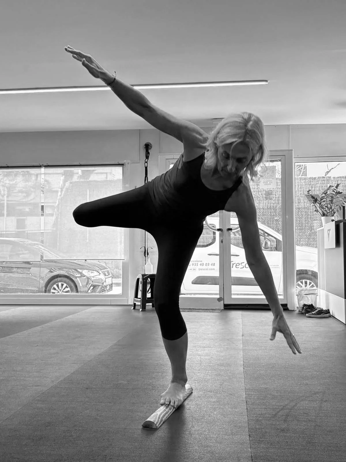Mujer haciendo yoga en un estudio, en posición de equilibrio sobre un pie con los brazos extendidos en diferentes direcciones, en un entorno con ventanas y autos afuera.