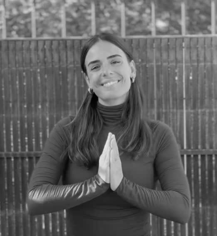 Mujer sonriendo con las manos juntas en forma de oración, de cabello largo, usa un suéter de manga larga y pendientes, y está frente a una cerca de madera.
