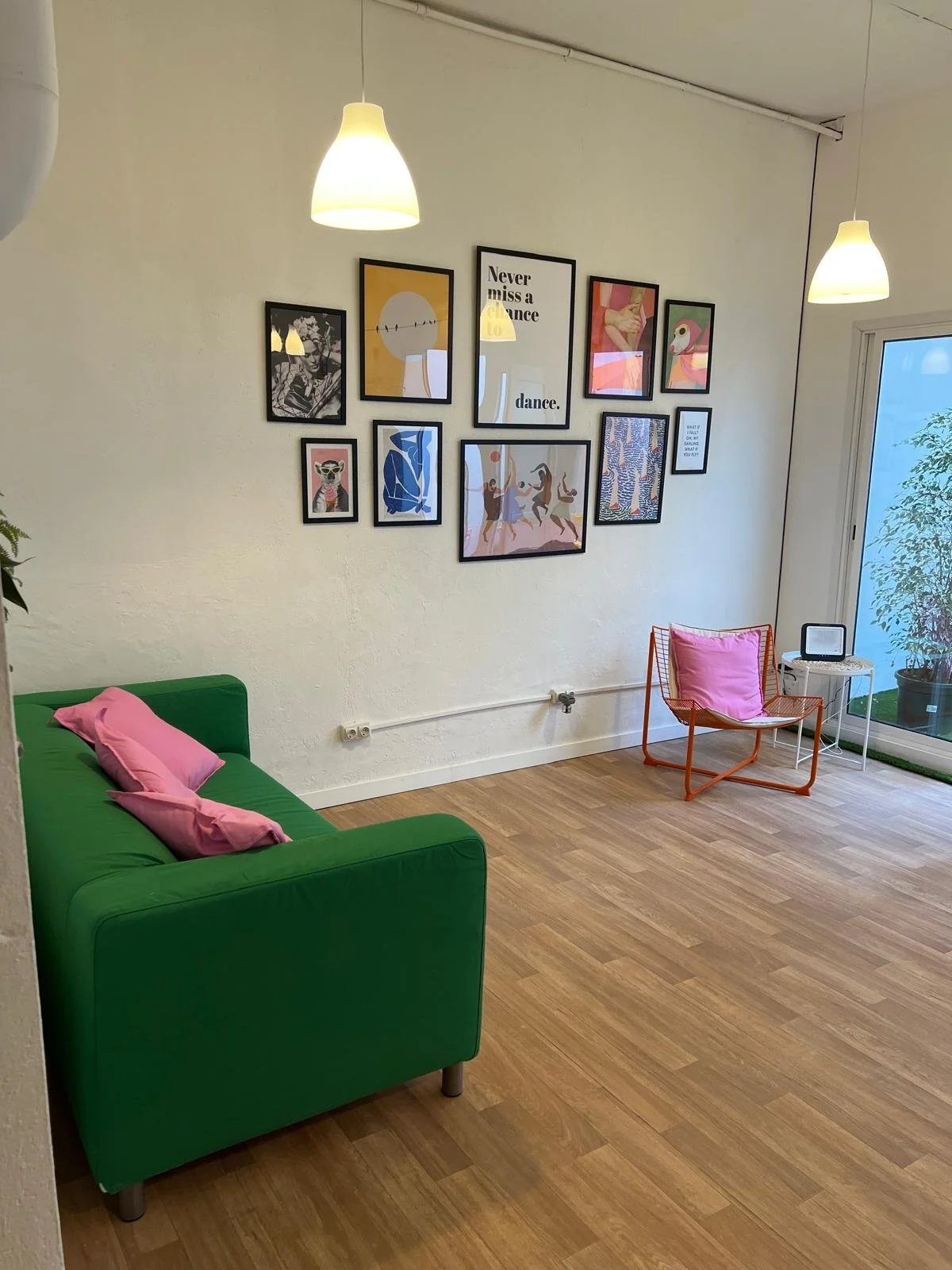 Salón con sillón verde, silla naranja con cojín rosado, pared decorada con cuadros y frases, lámparas colgantes, suelo de madera, ventana con balcón y planta