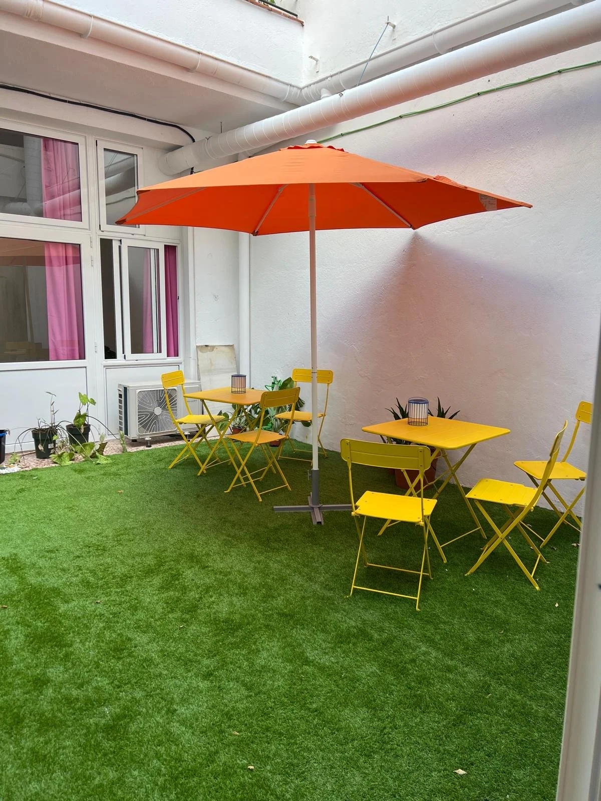 Espacio exterior con césped artificial, mesas y sillas amarillas, sombrilla naranja, plantas en macetas y ventanas con cortinas rosas.