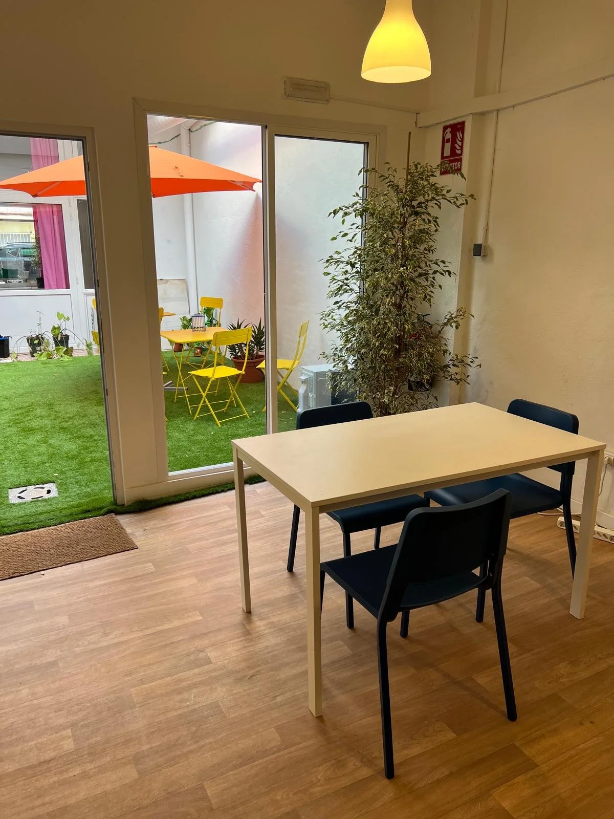 Interiores de una habitación con mesa y sillas negras y blancas, vista a un pequeño patio con césped artificial, mesa con sillas amarillas y un paraguas naranja.