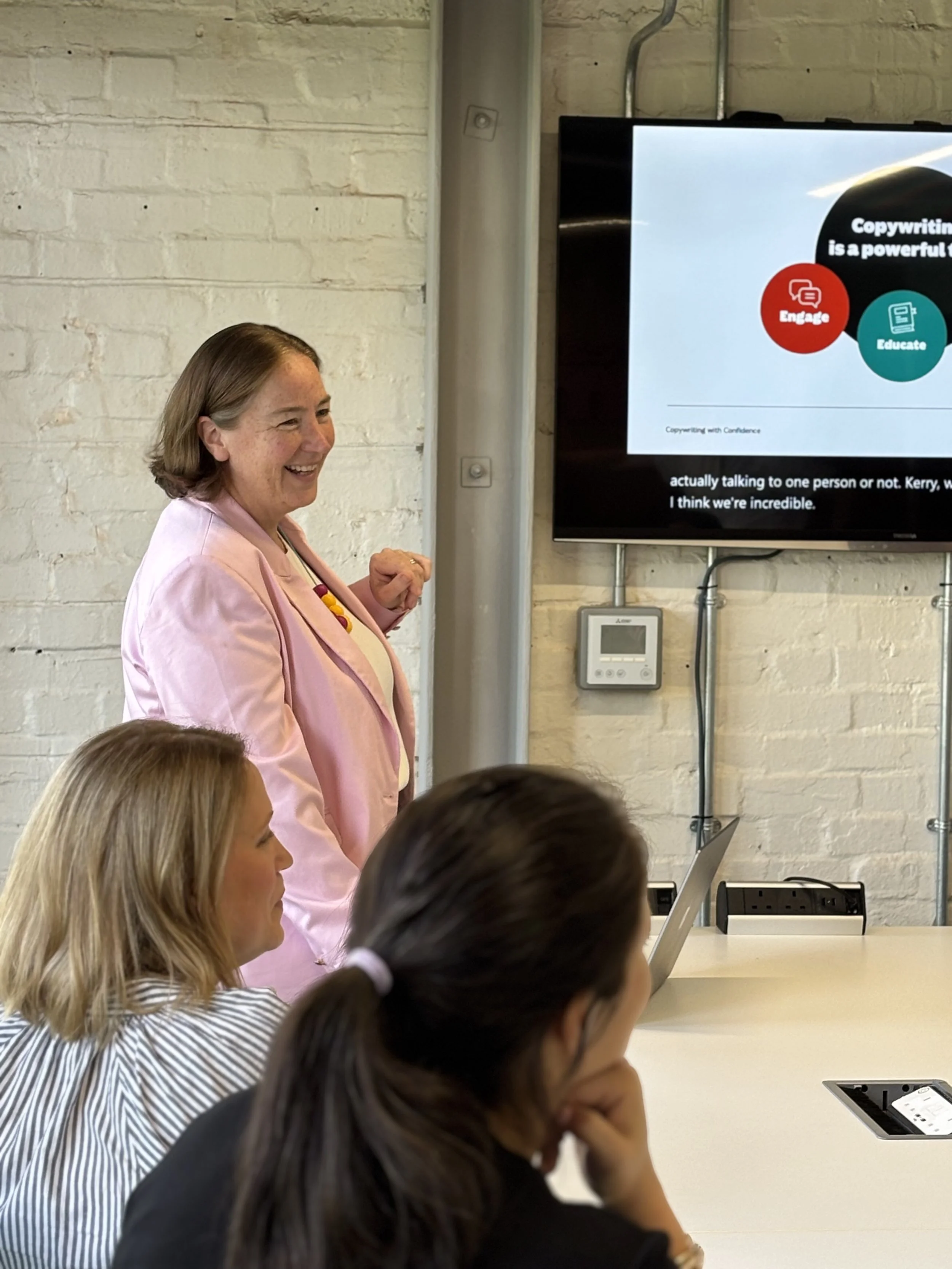 Sally Pritchett in a pink blazer giving a communications skills presentation, with a screen displaying a slide titled 'Copywriting is a powerful...'