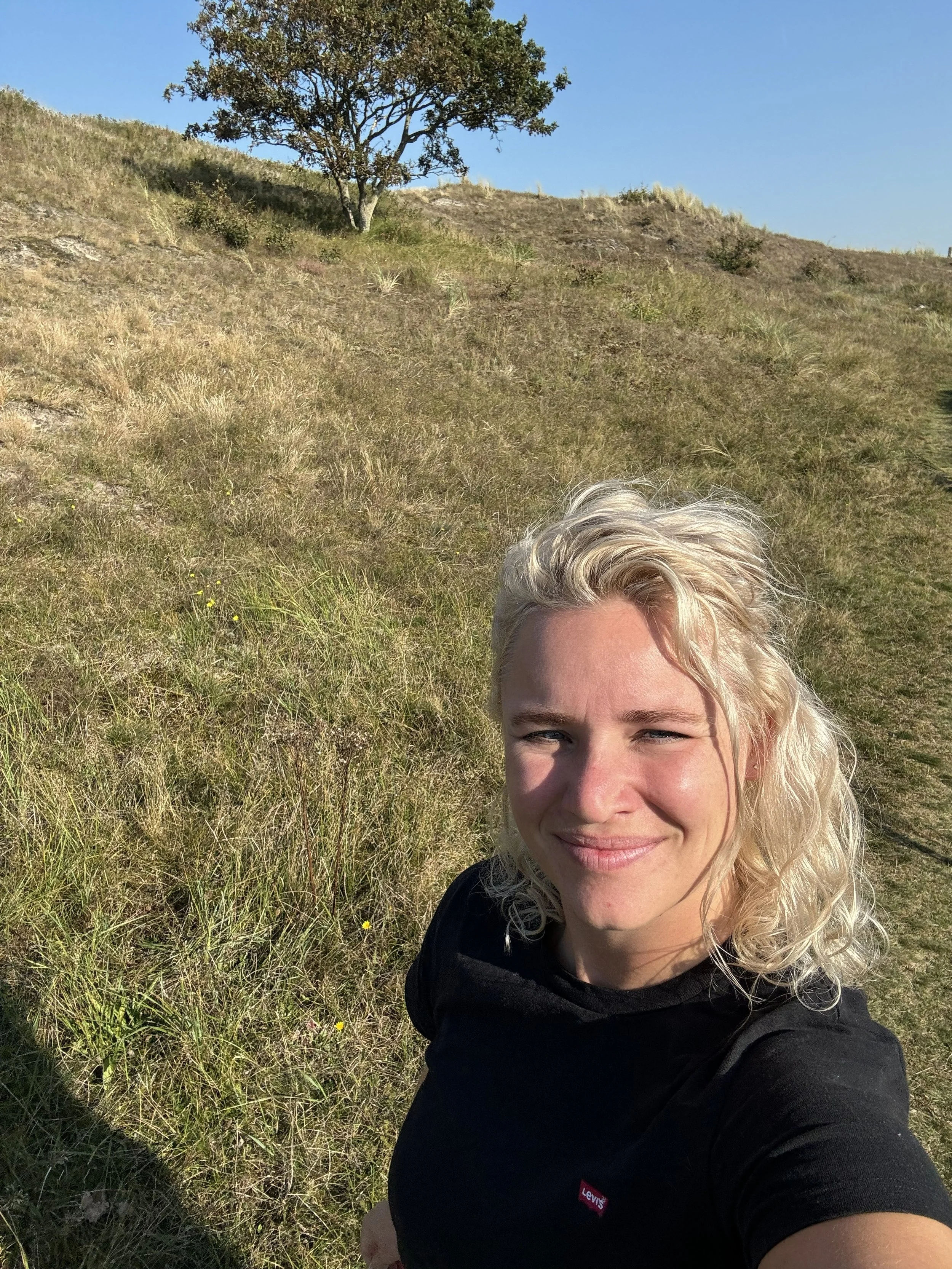 Een vrouw met blond krullend haar die een glimlach heeft, neemt een selfie in een grasrijk, heuvelachtig landschap onder een blauwe lucht, met een enkele boom op de achtergrond.