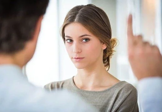 A young woman with brown hair and a gray shirt listens carefully during a conversation with two people whose faces are not visible.