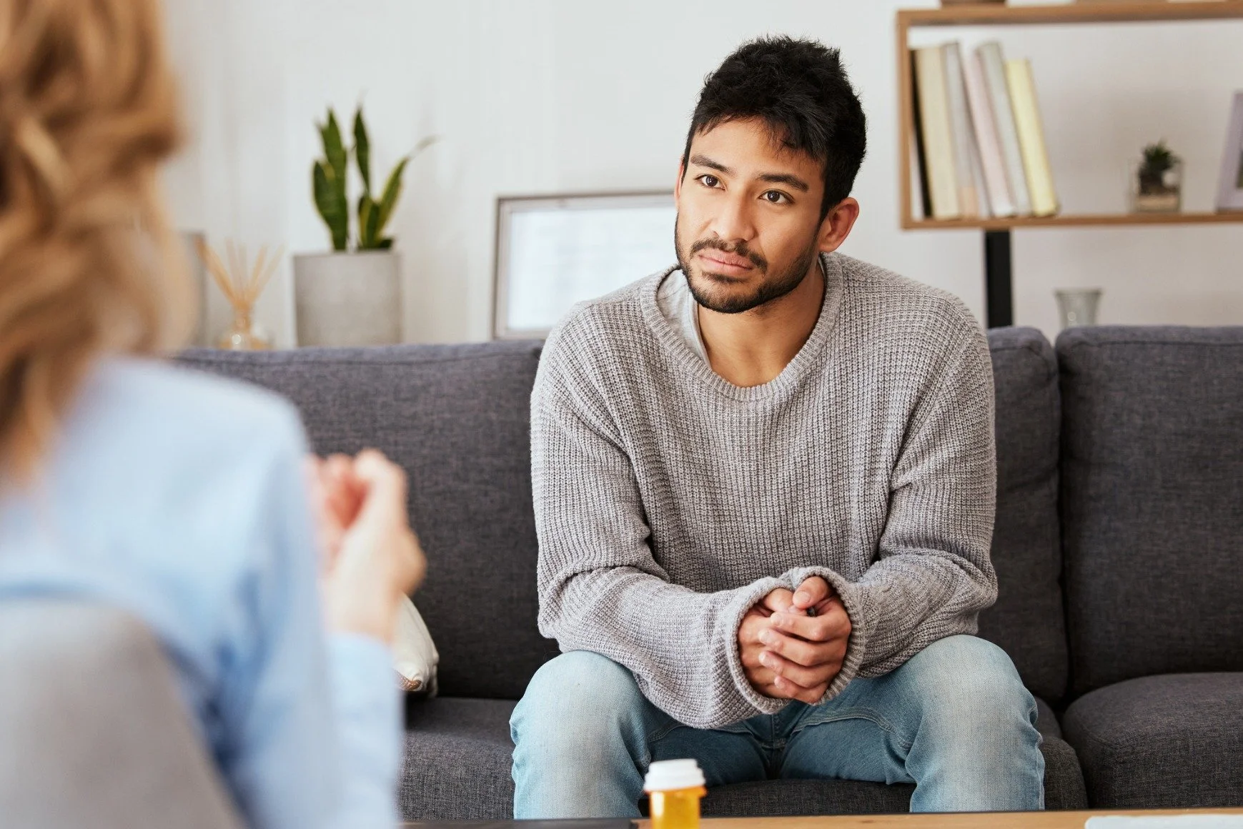 A man sitting on a gray sofa, having a conversation with a woman whose back is facing the camera. The background includes a bookshelf and plants.