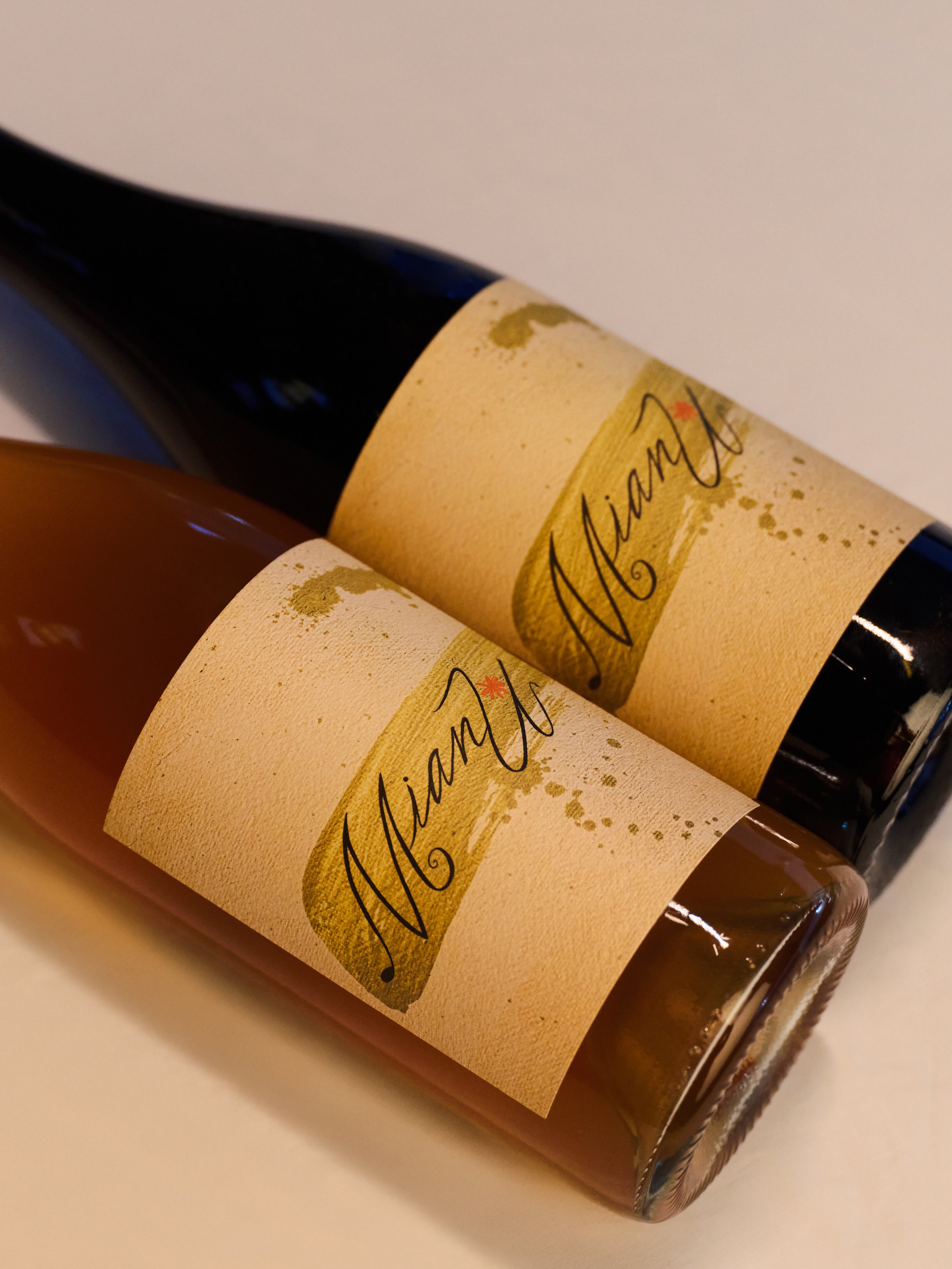 Two bottles of MianU Holiday Syrah and MianU Orange wine lying on their sides, one orange and one red, with beige labels that have the word 'MianU' written in black cursive.