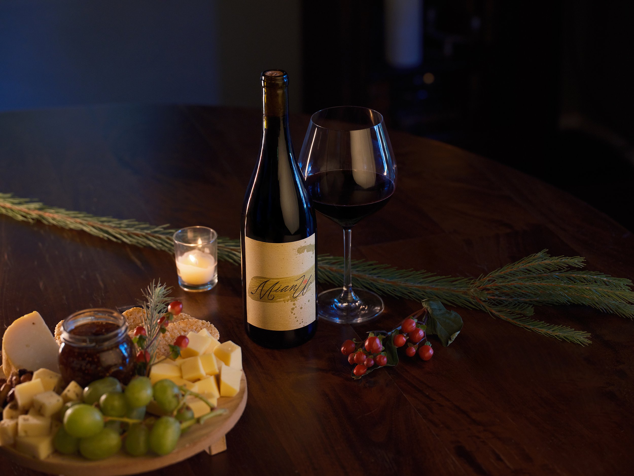 A bottle of MianU Holiday Syrah, a glass of red wine, a small candle, a cheese platter with grapes, cheese, chocolate, and crackers, and holiday greenery and berries on a dark wooden table.