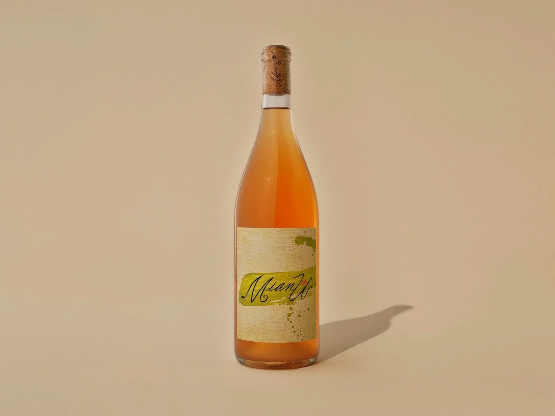 A bottle of MianU Orange wine with a beige, textured label that has green paint splashes and a handwritten-style "MianU" on it. The bottle has a cork seal with some cursive writing.