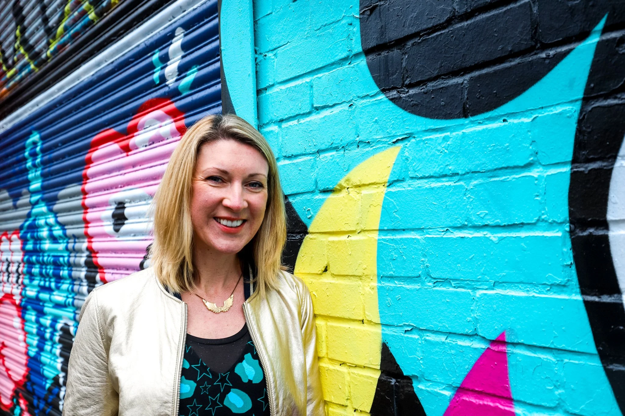 Ashleigh standing in front of a colourful street mural painted on a brick wall.