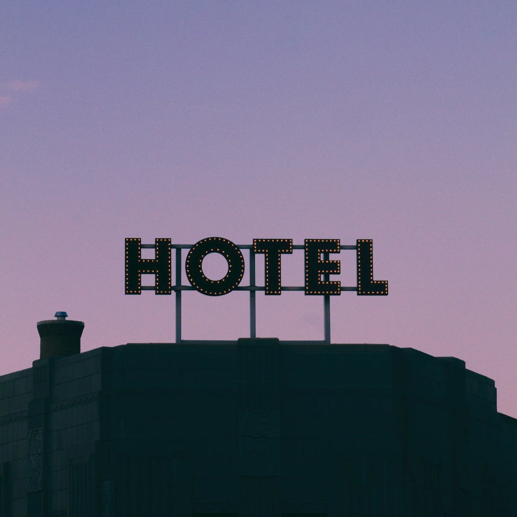 An illuminated hotel sign on top of a dark building with a purple sky background.